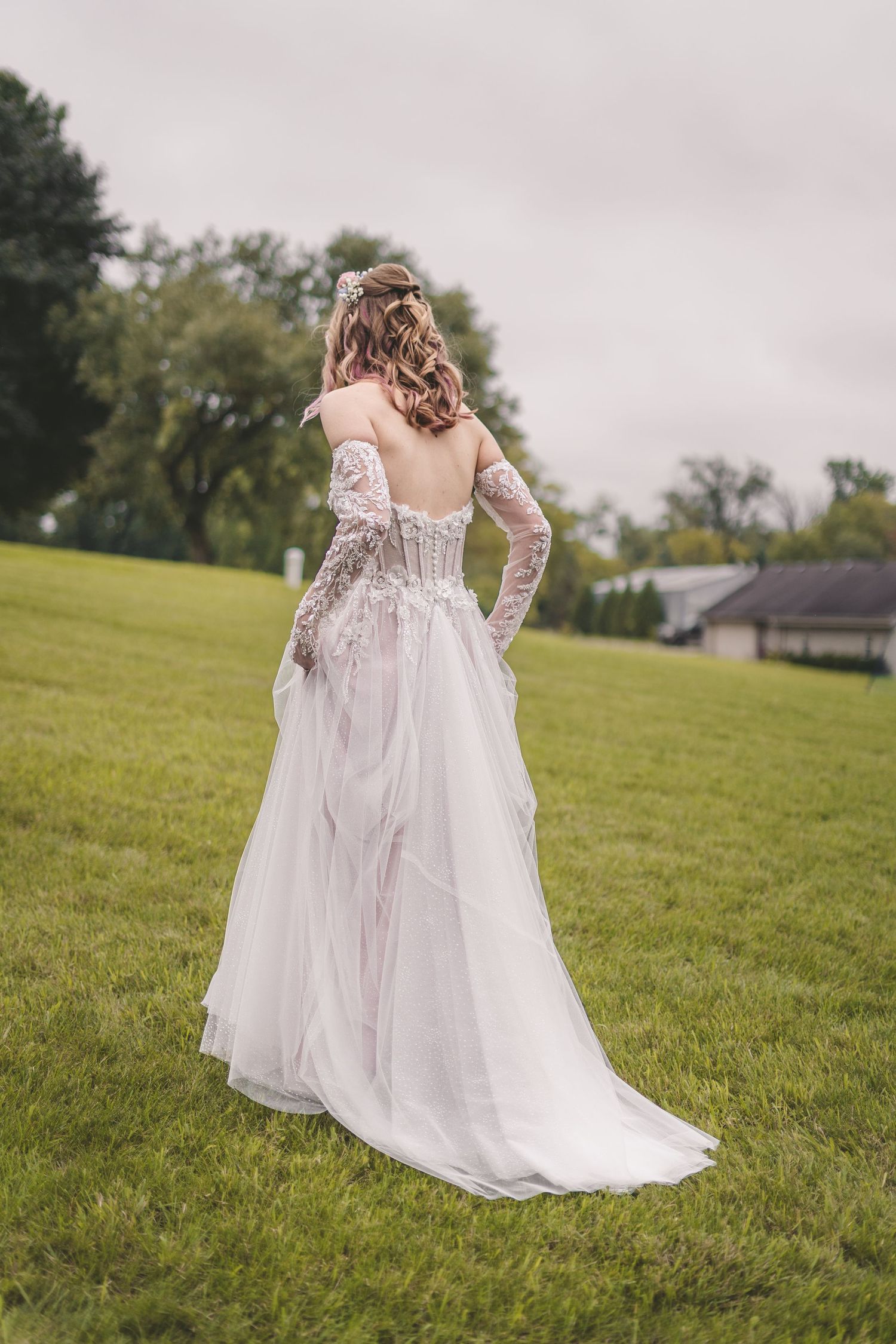 Bride in elegant off-shoulder lace wedding gown with tulle skirt standing on green lawn, back view.