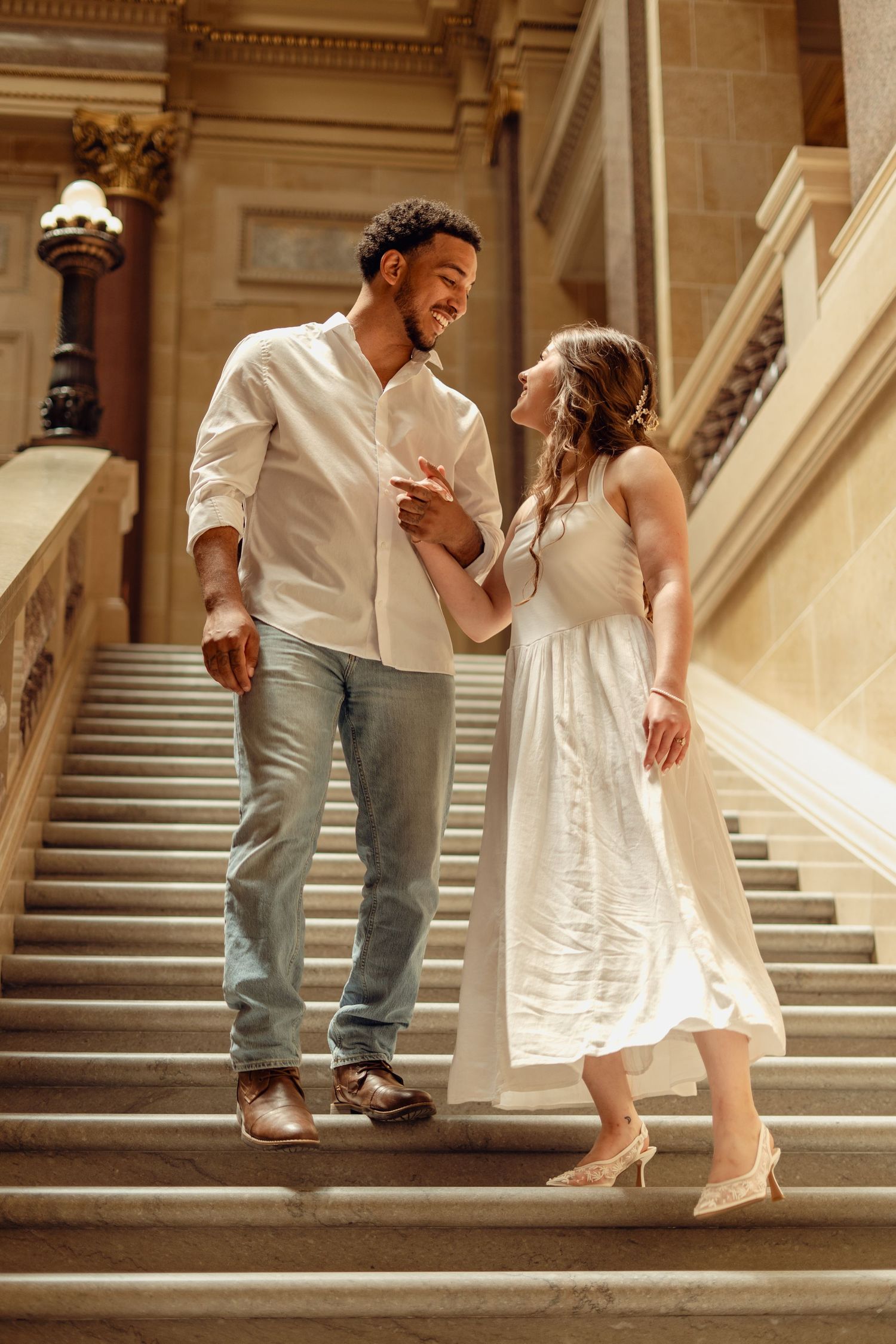 Couple in white outfits smiling at each other on grand marble staircase in elegant building.