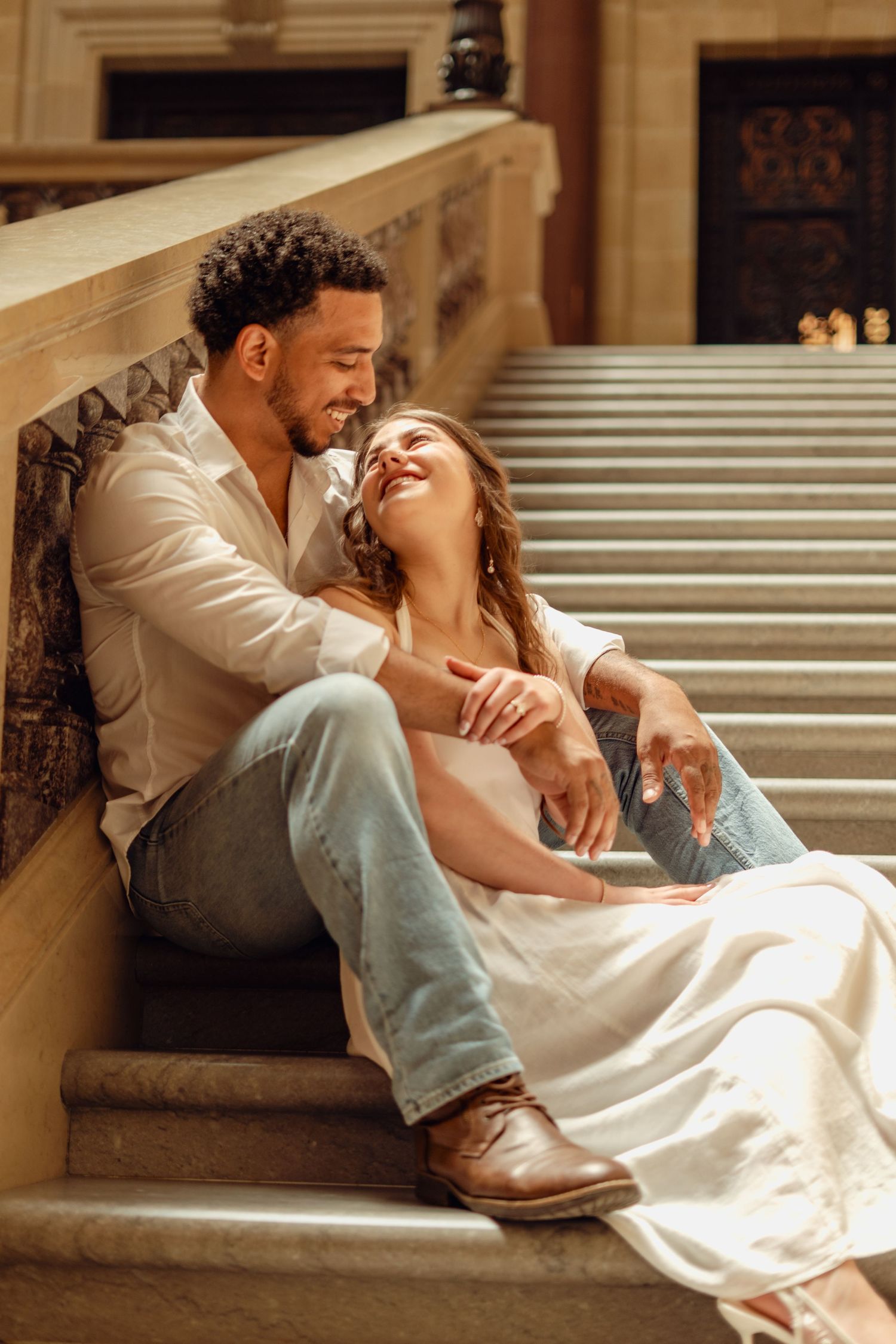 Romantic interracial couple sitting on marble stairs, woman leaning against man in white shirt and jeans, smiling.