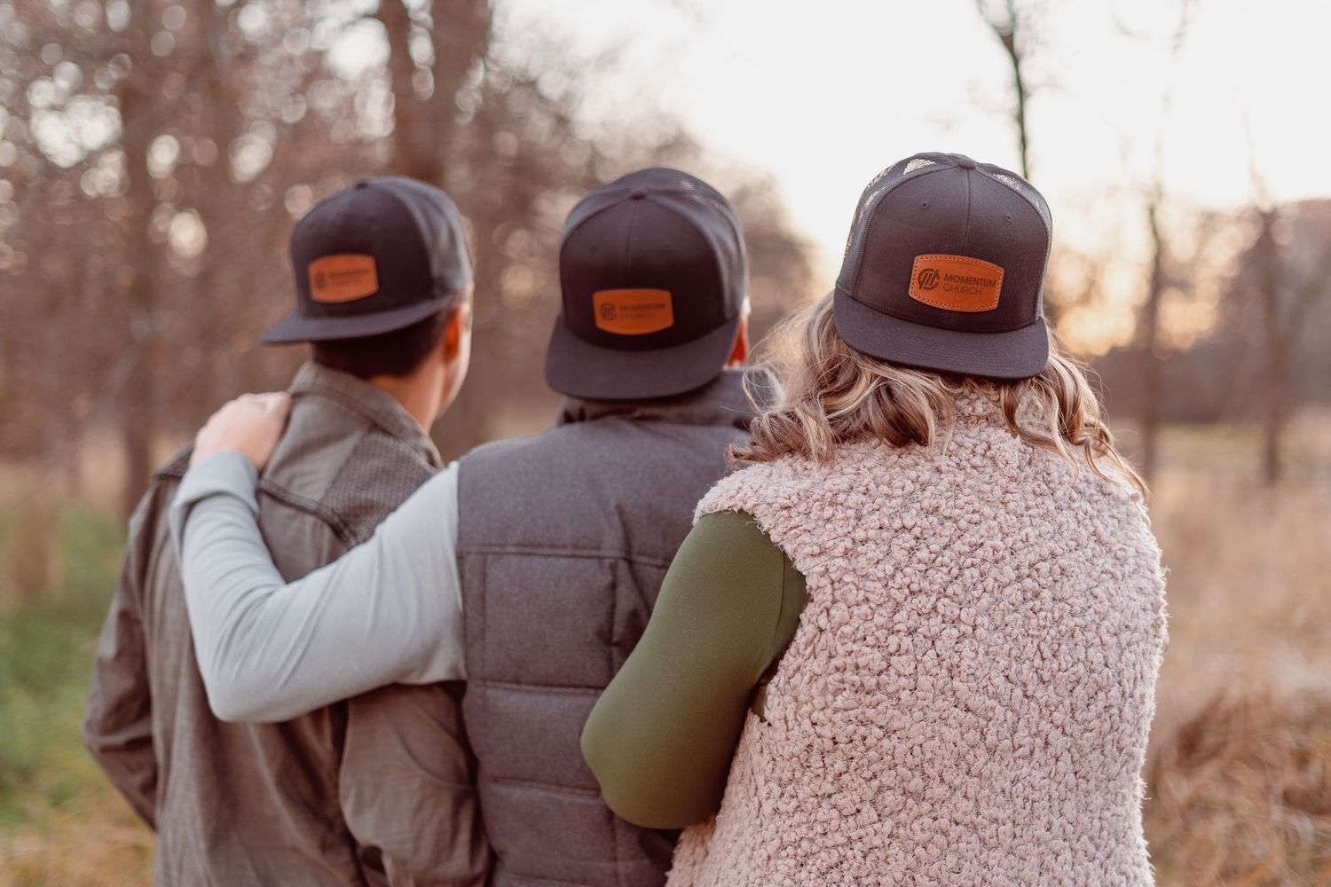 Three people wearing matching brown trucker hats with leather patches stand together outdoors in a wooded autumn setting.