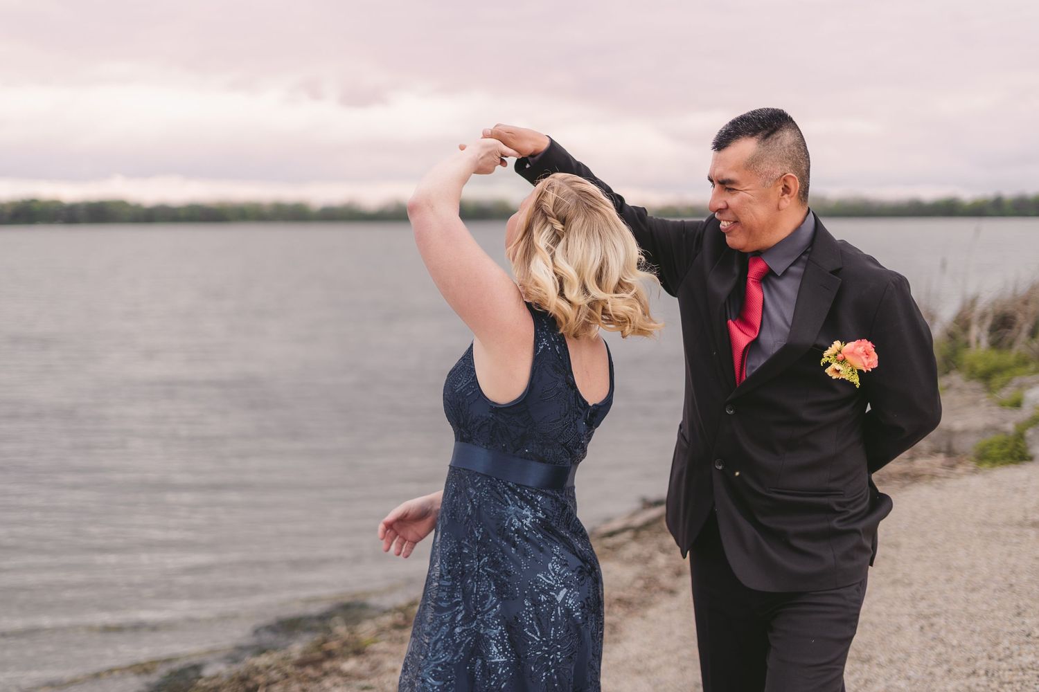 Couple dancing by the waterfront, woman in blue sequin dress being twirled by man in black suit with red tie.
