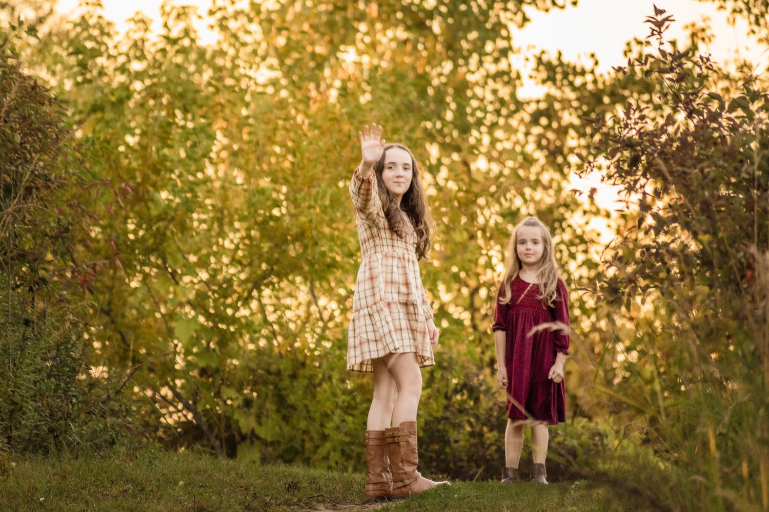 Two children in dresses stand in a golden-lit field during sunset.