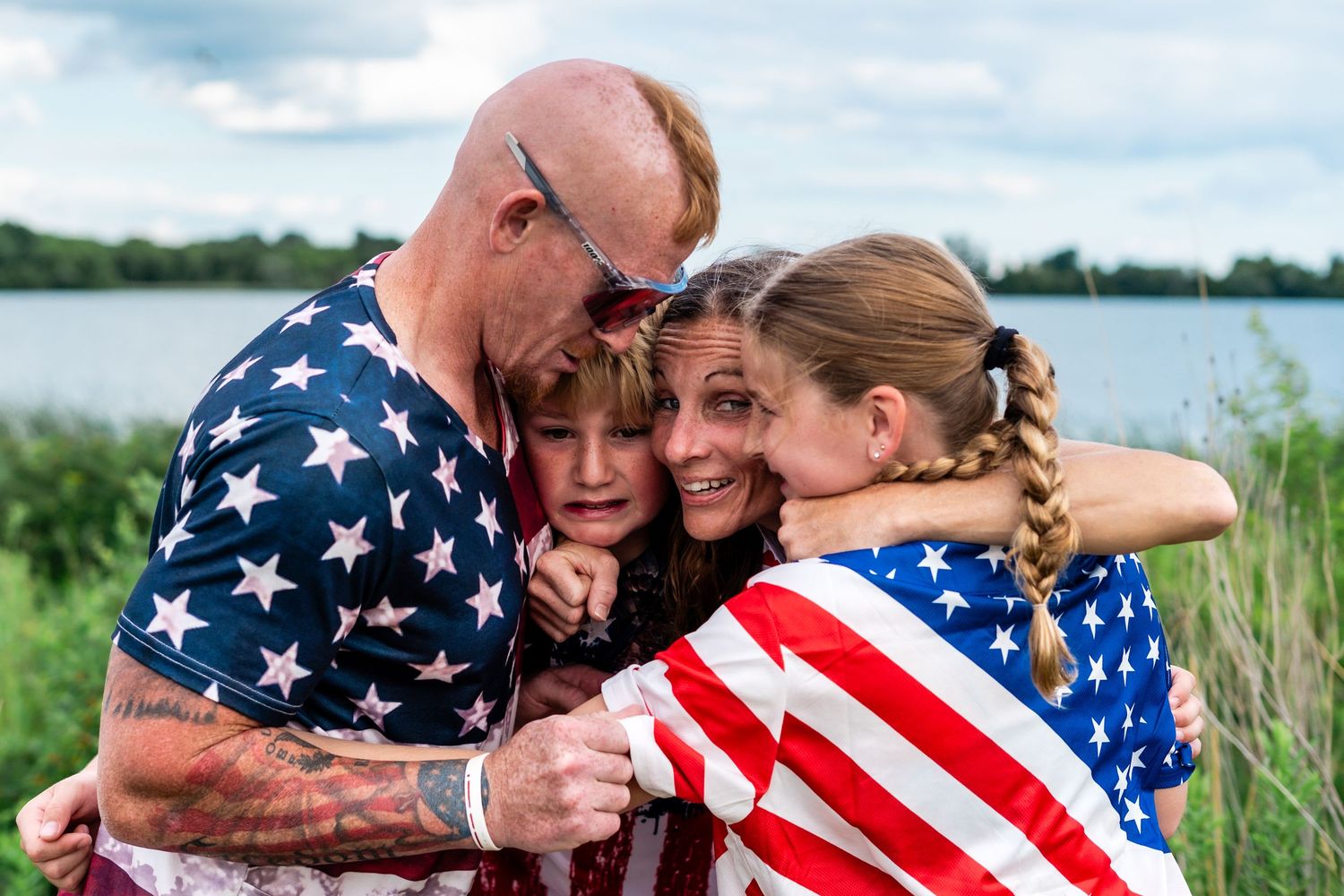 Family in American flag attire sharing a warm group hug outdoors near a lake on a patriotic occasion.