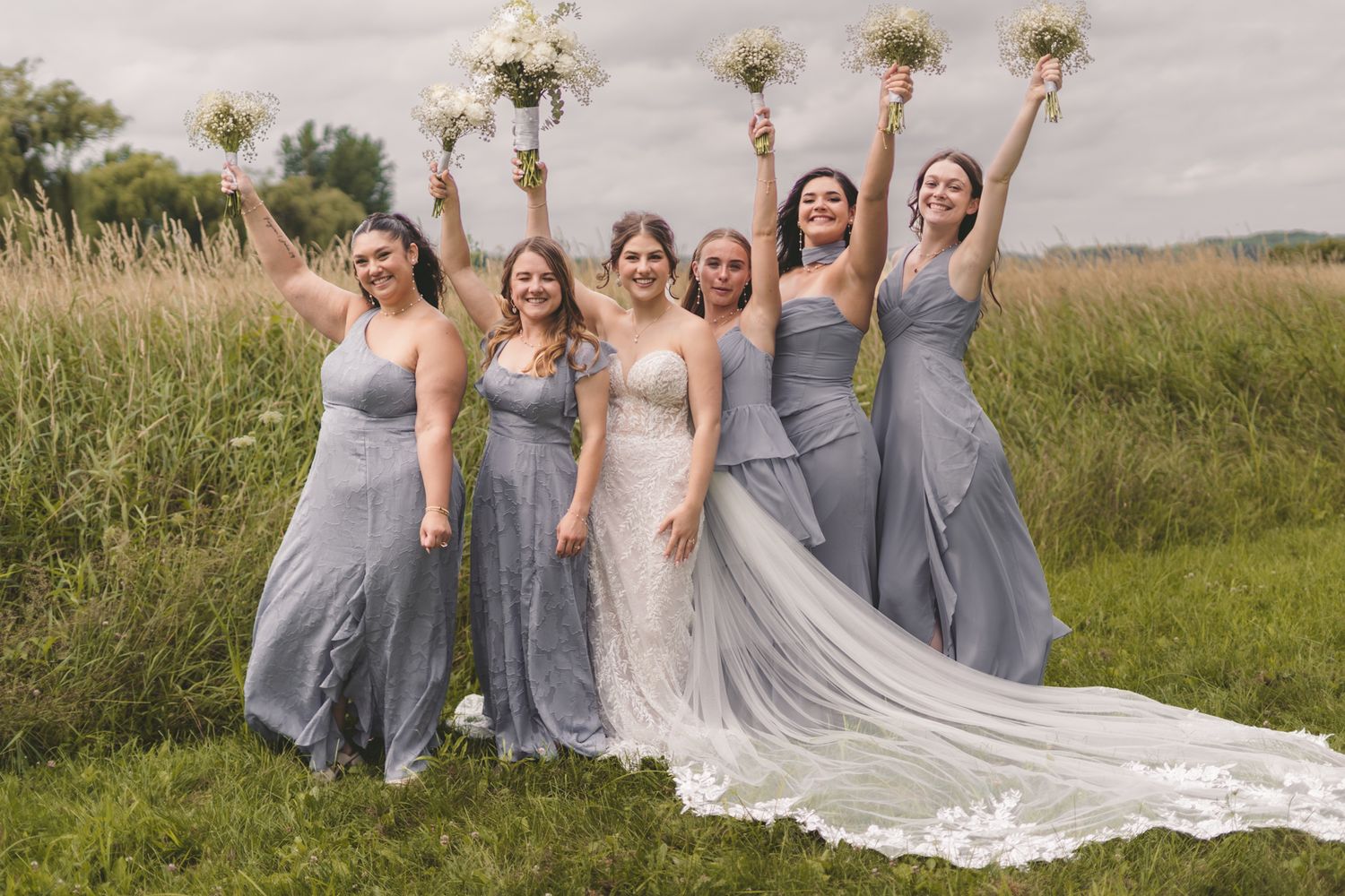 Bride in white lace gown with five bridesmaids in blue-gray dresses celebrating outdoors in a field.