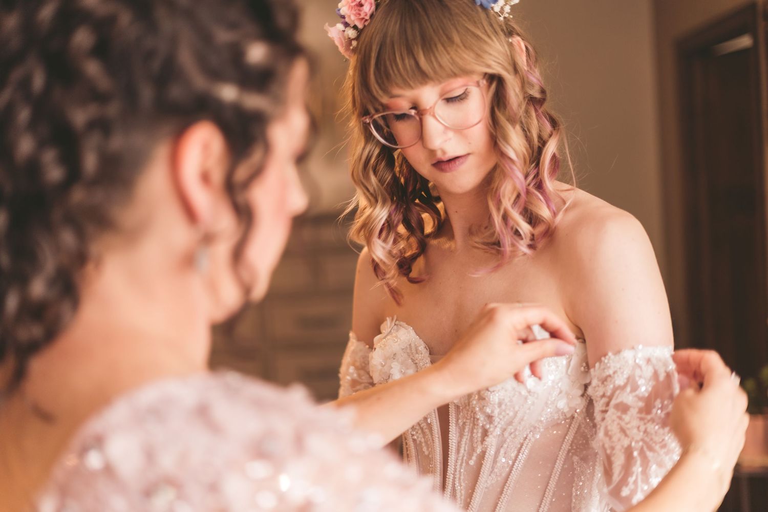 Bridesmaid helping bride adjust her lace off-shoulder wedding dress while bride wears floral hair accessories.