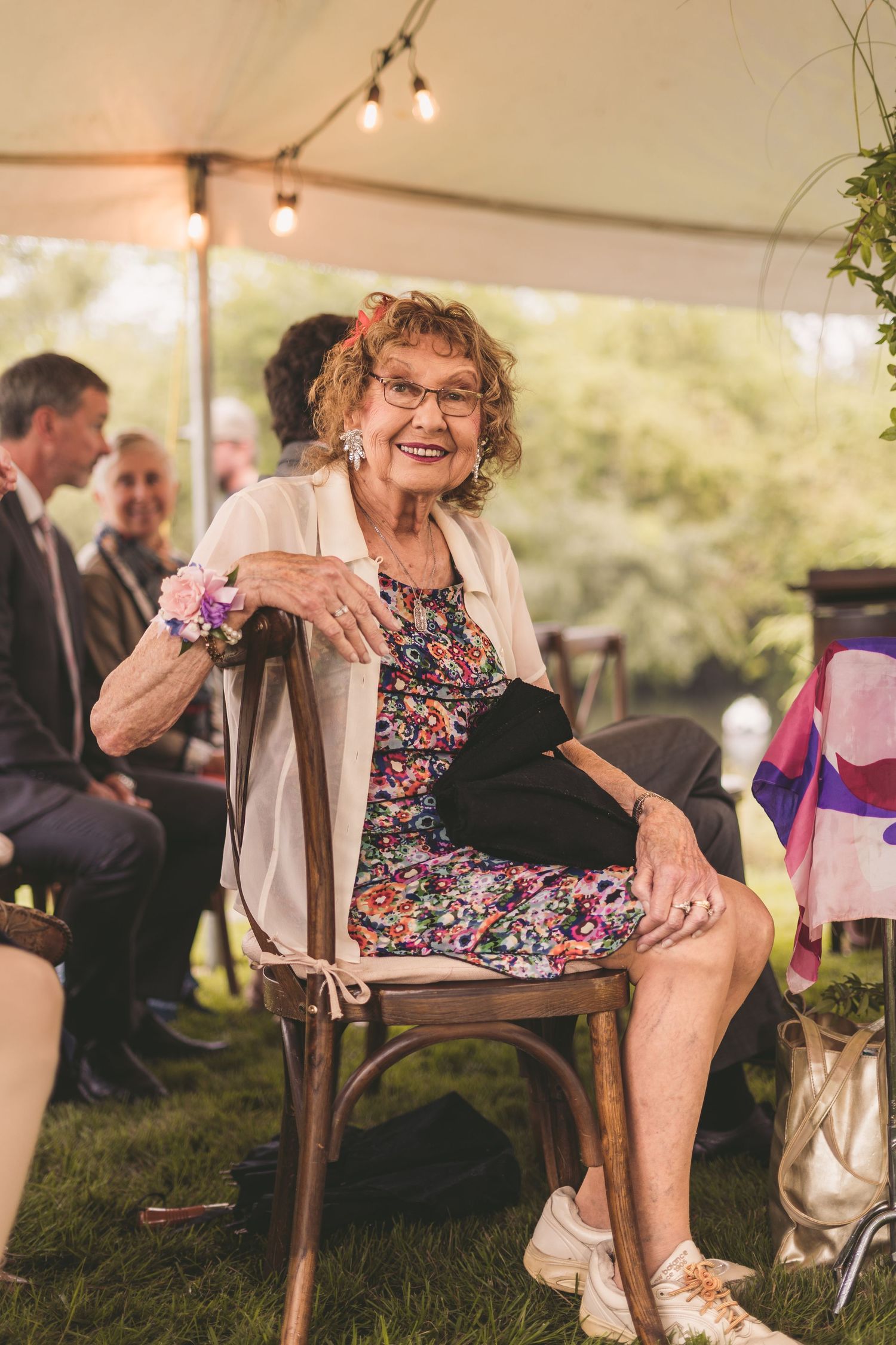 Elderly woman smiling in floral dress with wrist corsage seated at outdoor wedding.