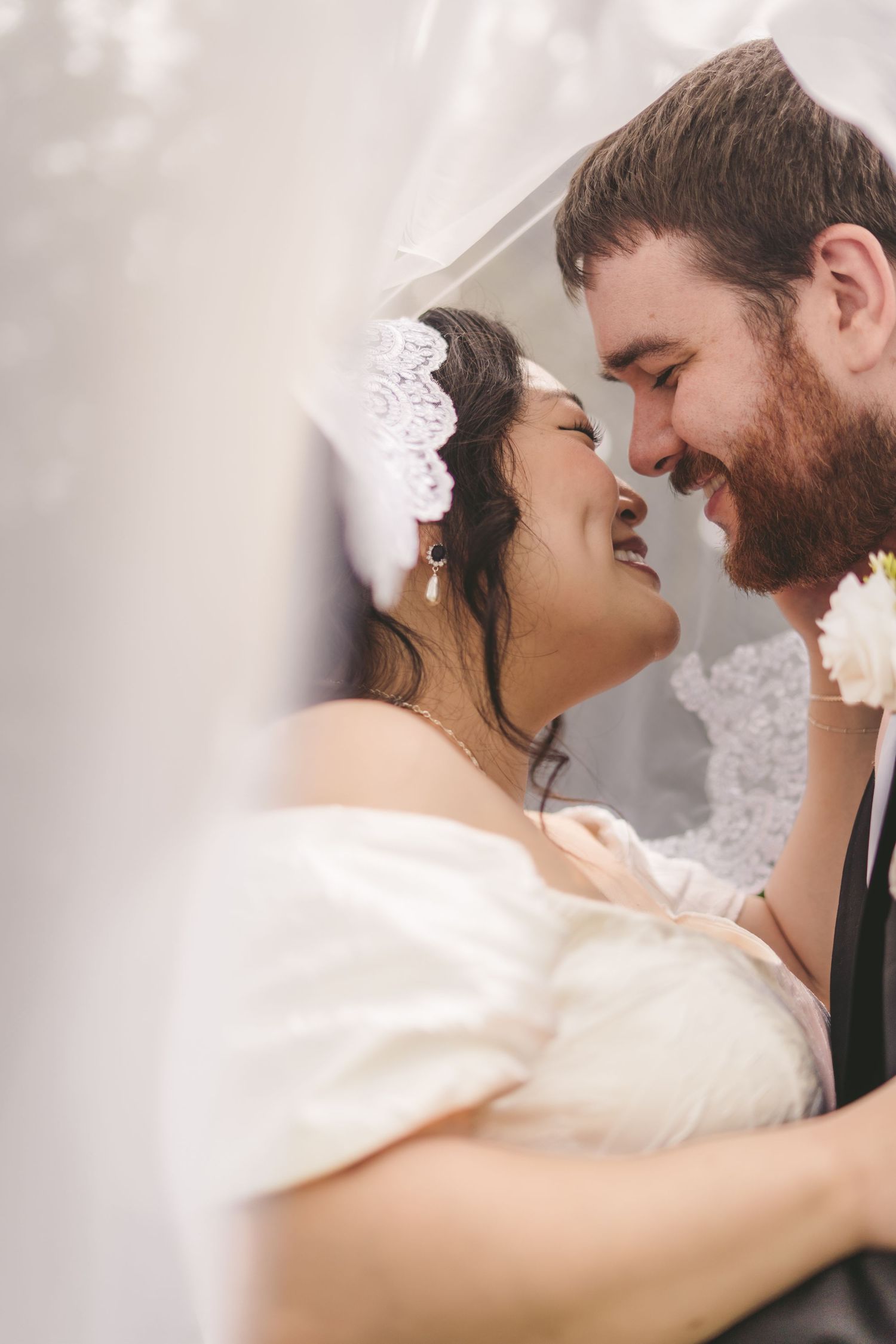 A smiling bride and groom share an intimate moment under a wedding veil, the groom with a beard and the bride wearing a lace headpiece.