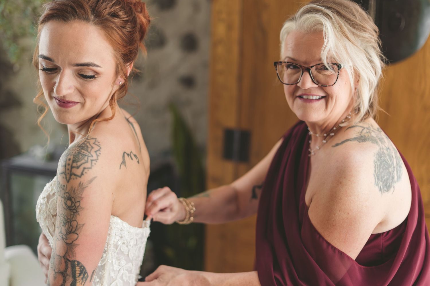 Bride in white lace gown with tattoos gets dress fastened by older woman in burgundy dress, both smiling.