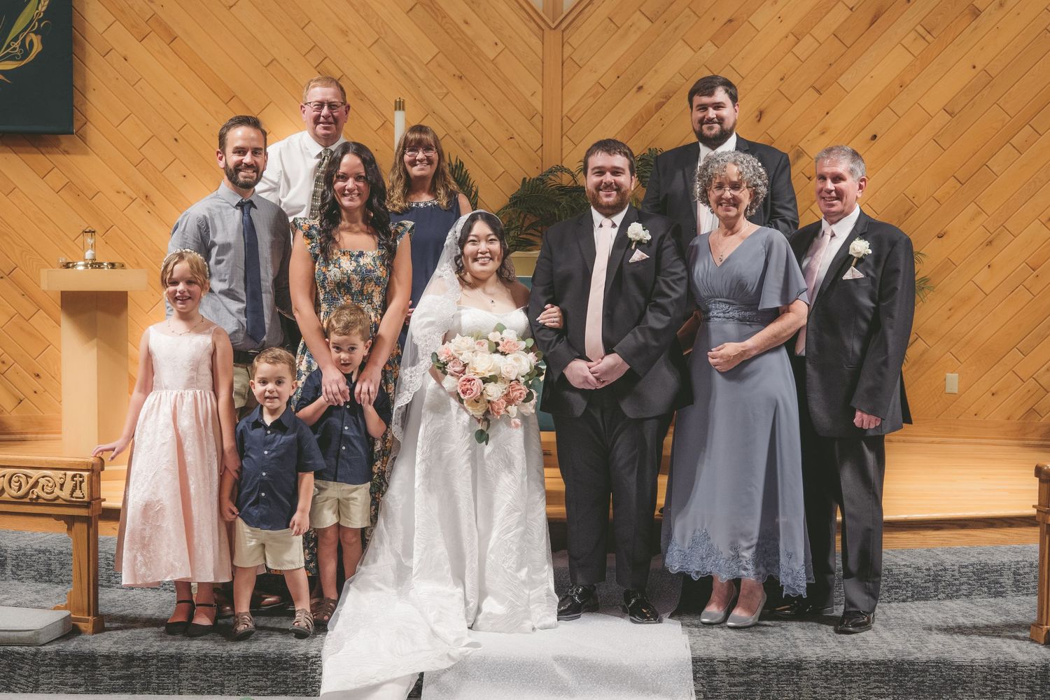 Wedding party poses together in a wood-paneled church, bride in white gown holding bouquet surrounded by family.