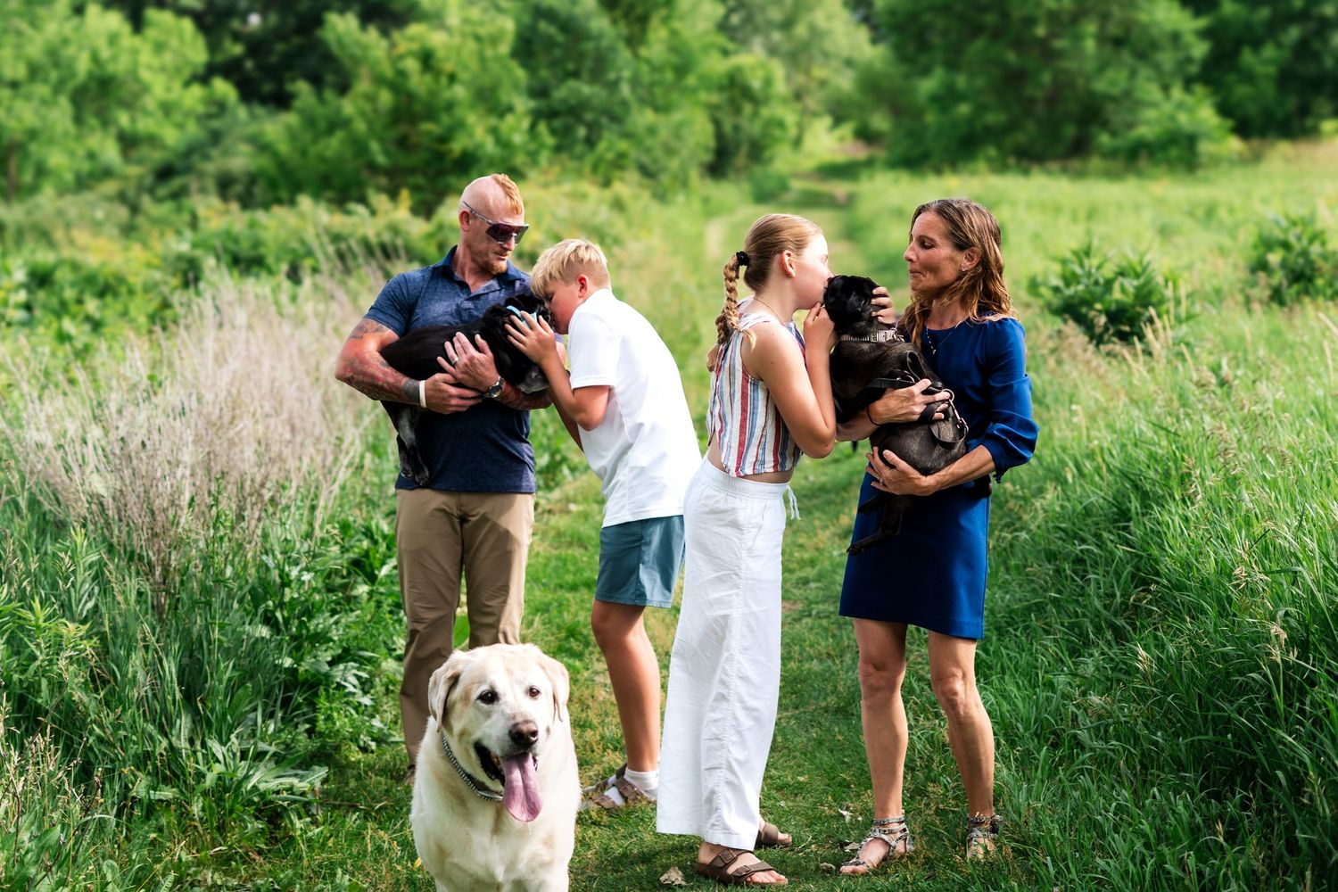 Family with two small dogs and a white Labrador on a lush green nature trail during an outdoor photo session.