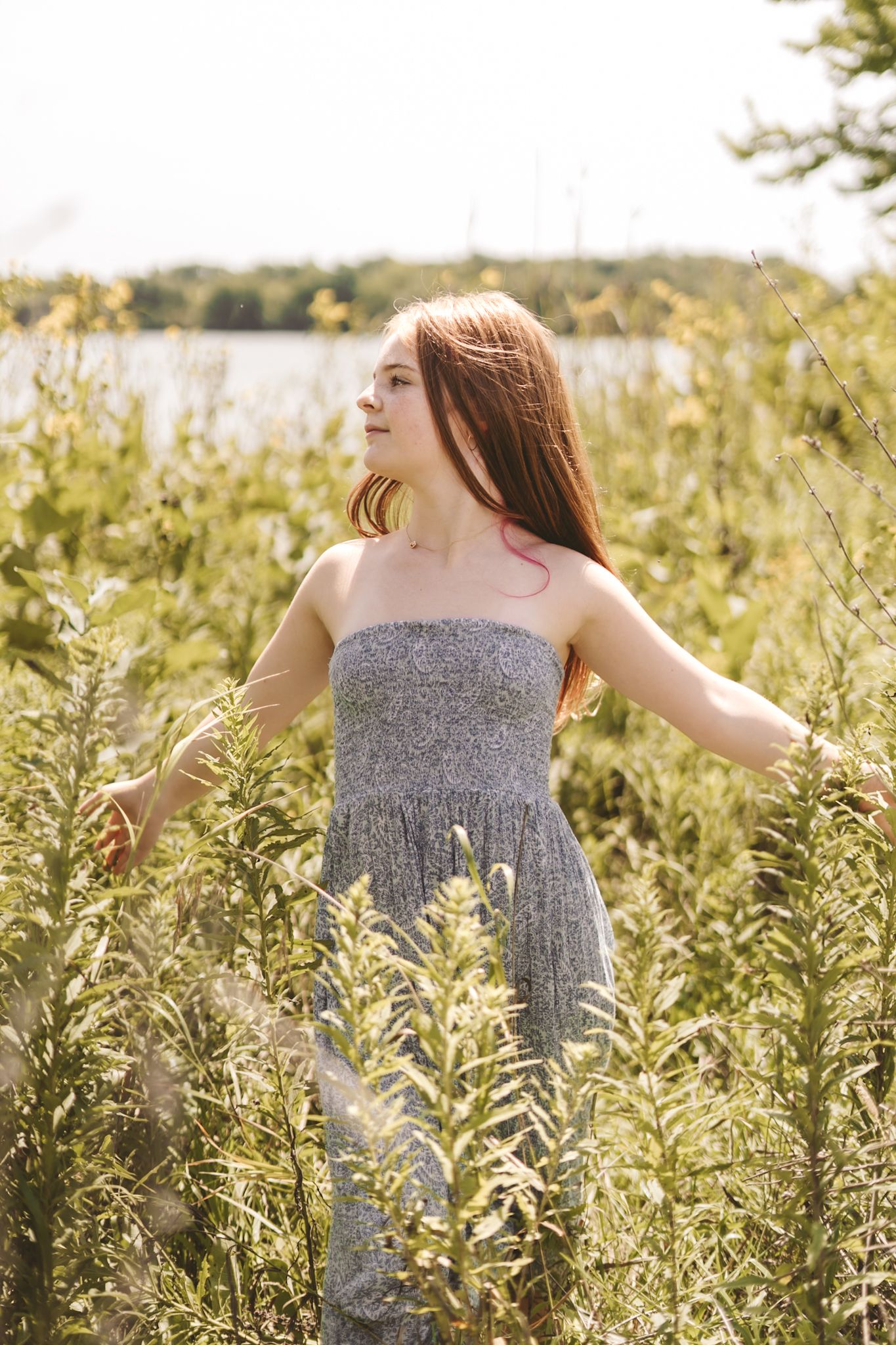 Young redhead woman in gray strapless dress standing among tall green wildflowers near a lake on a sunny day.