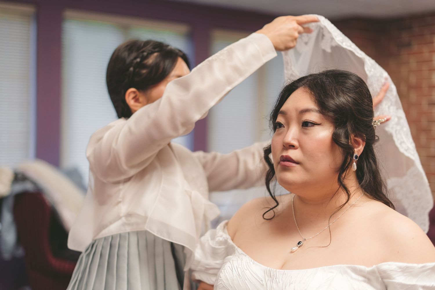 A bride in a white dress has her veil adjusted by a bridesmaid in a sheer top during wedding preparations.