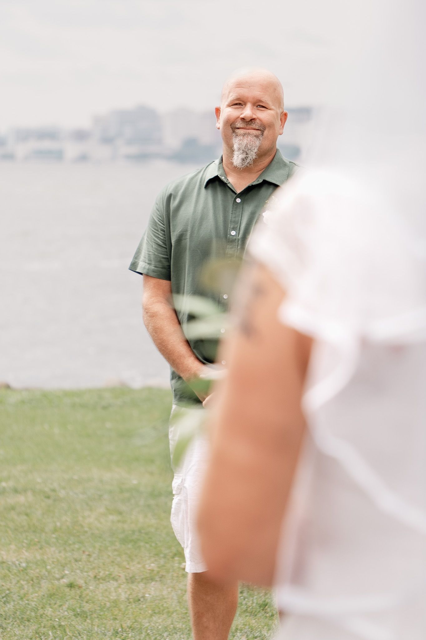 A person in green polo and white shorts stands on grass near water.