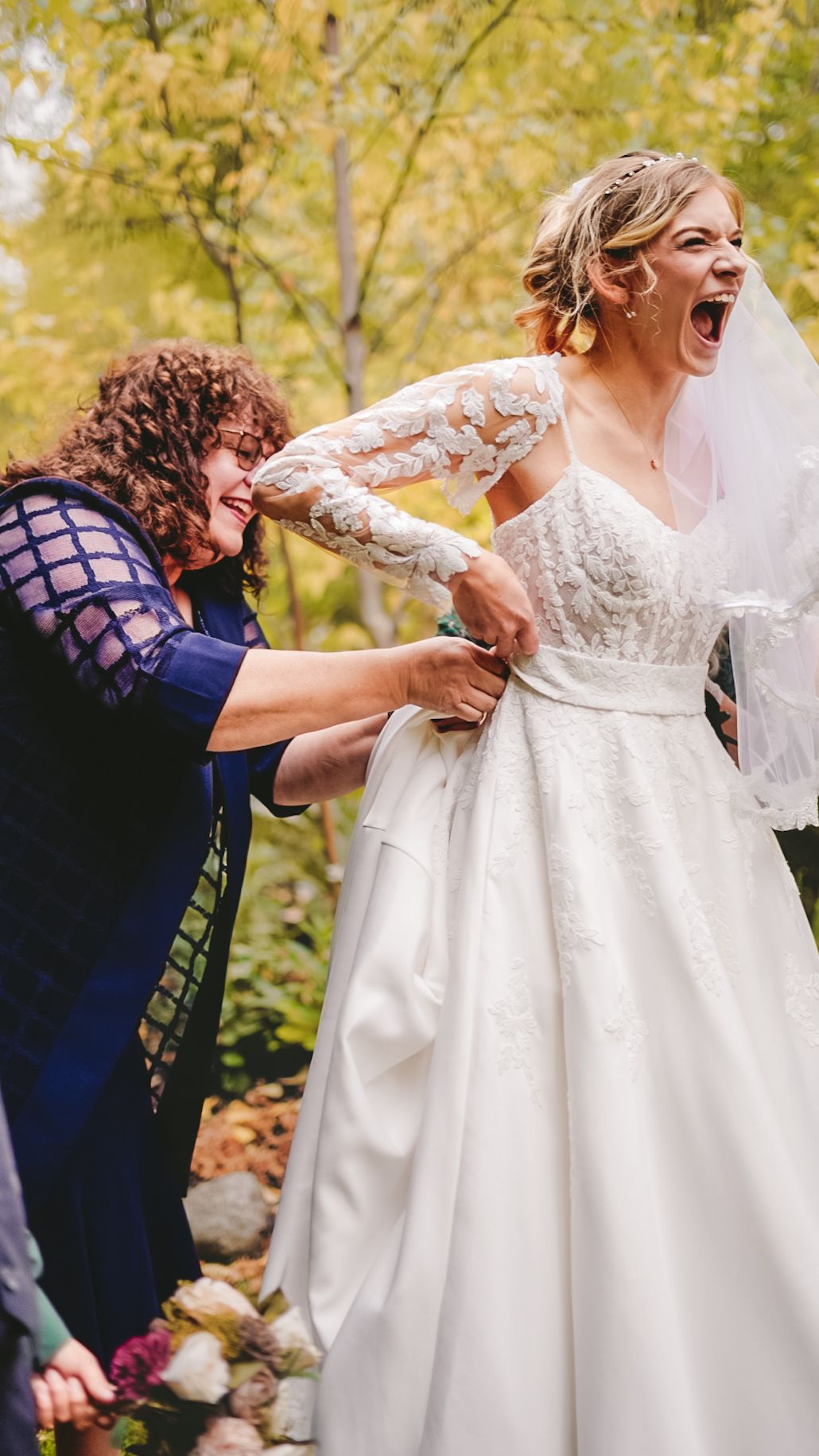 Bridesmaids help adjust a white lace wedding dress outdoors.