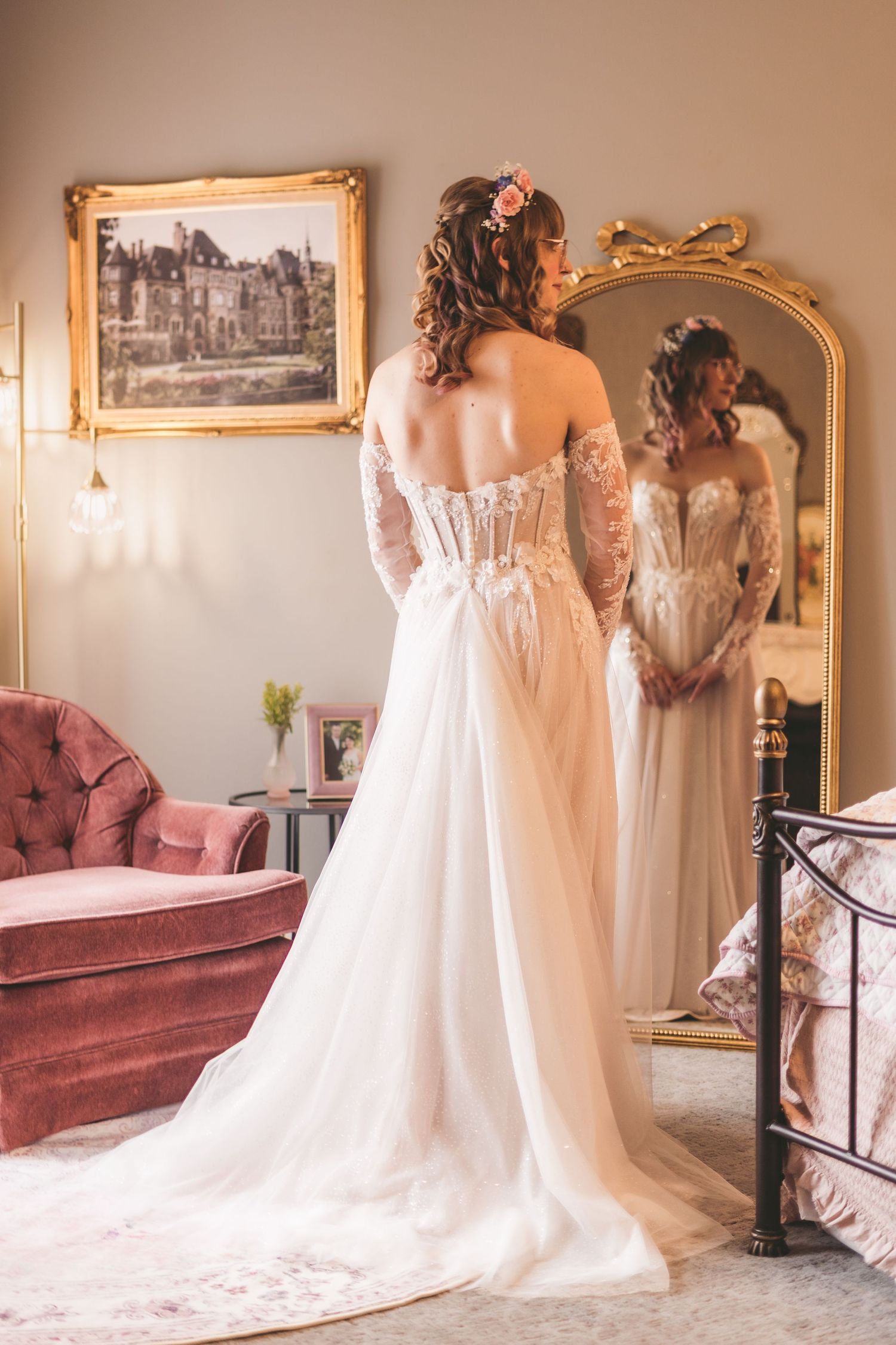 Bride in elegant lace wedding gown with corset back and floral hair accessory standing before ornate gold mirror.
