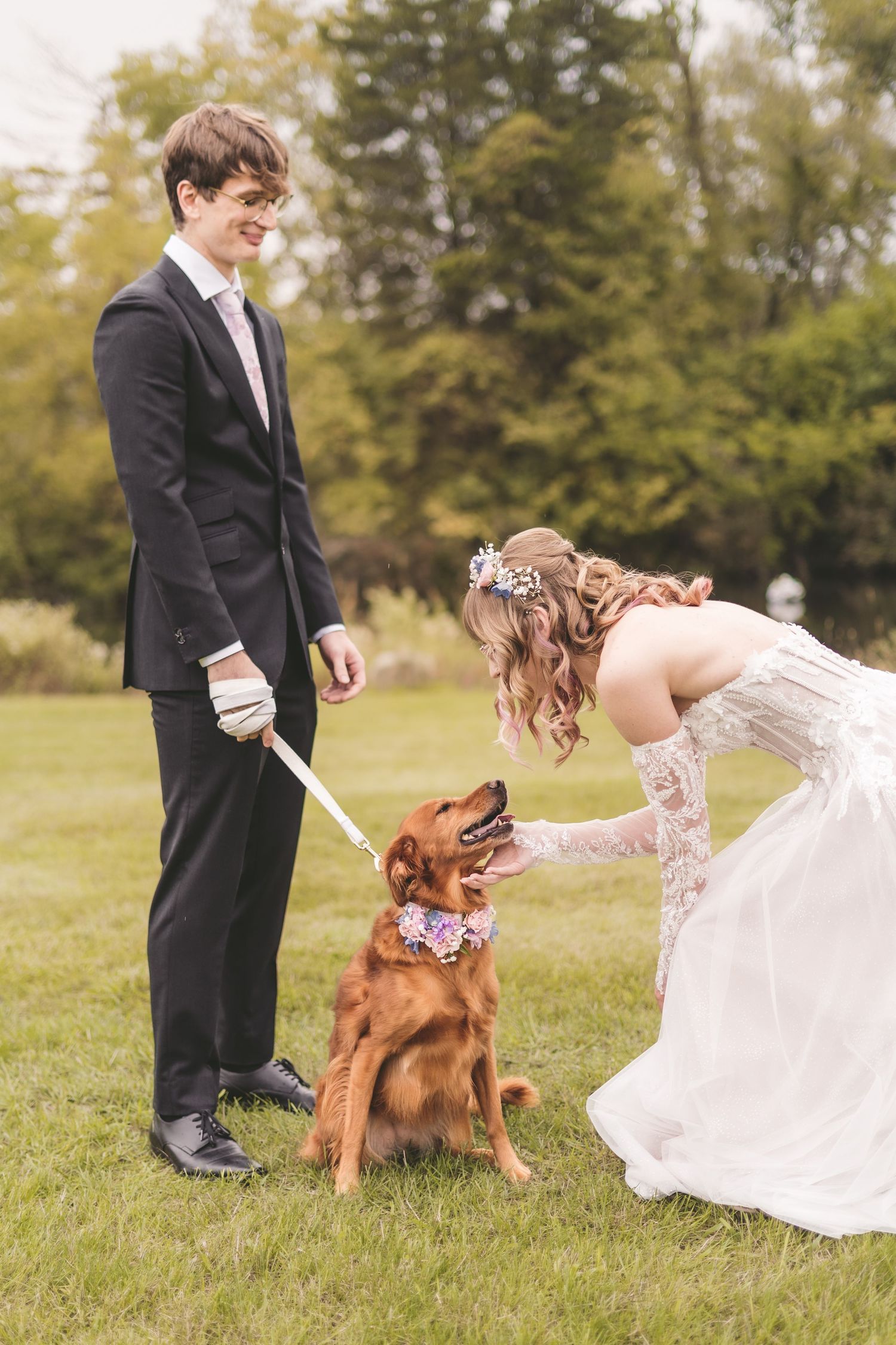 Bride and groom with their golden retriever dog wearing floral collar at outdoor wedding.