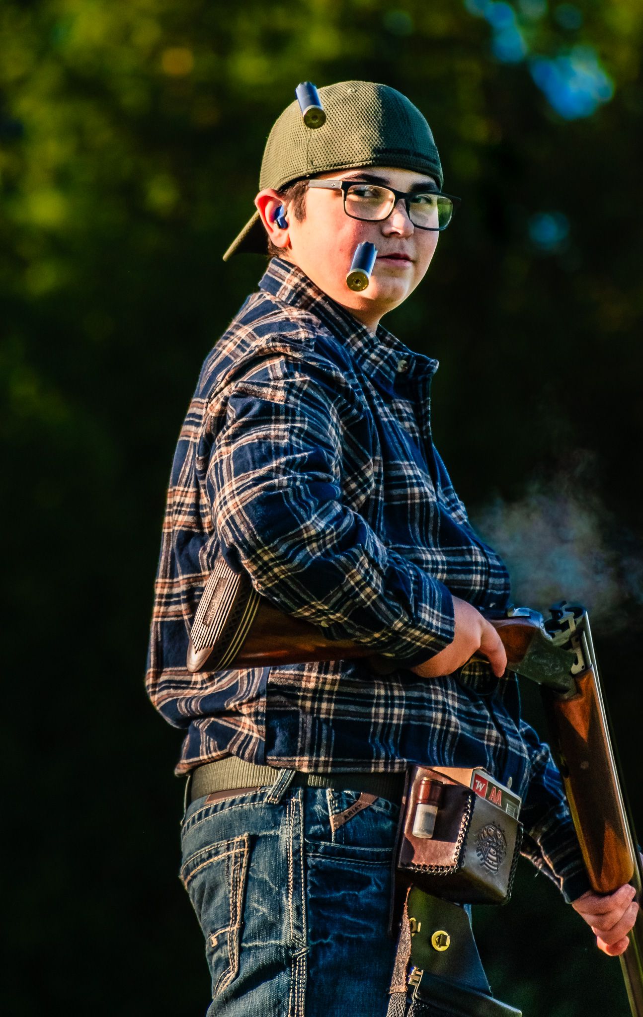 Person in plaid shirt and baseball cap holding hunting equipment outdoors.
