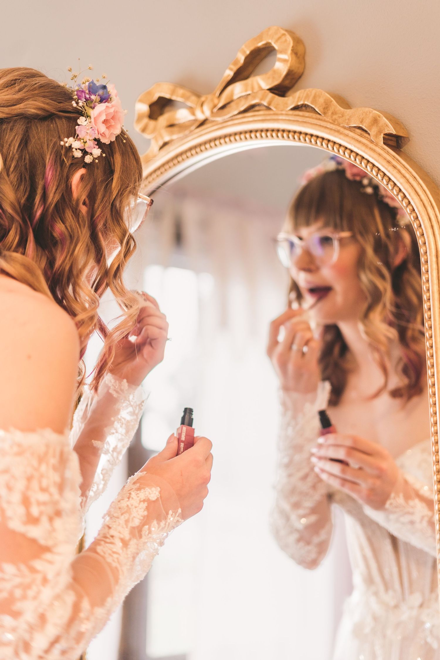 Bride with floral hair accessories applying lipstick while looking into an ornate gold mirror in bridal gown.