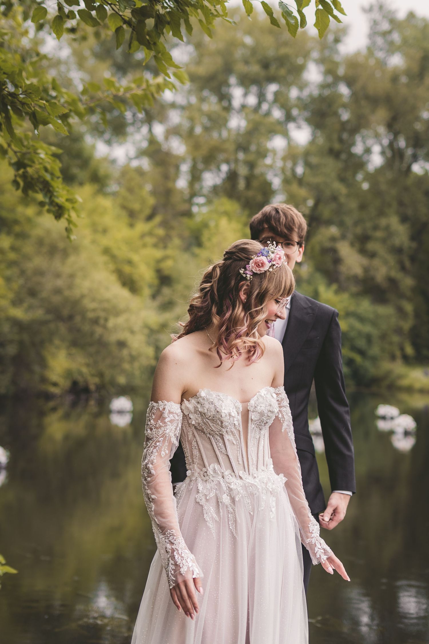 Bride in lace corset wedding dress with groom in black suit standing by a serene forest pond.