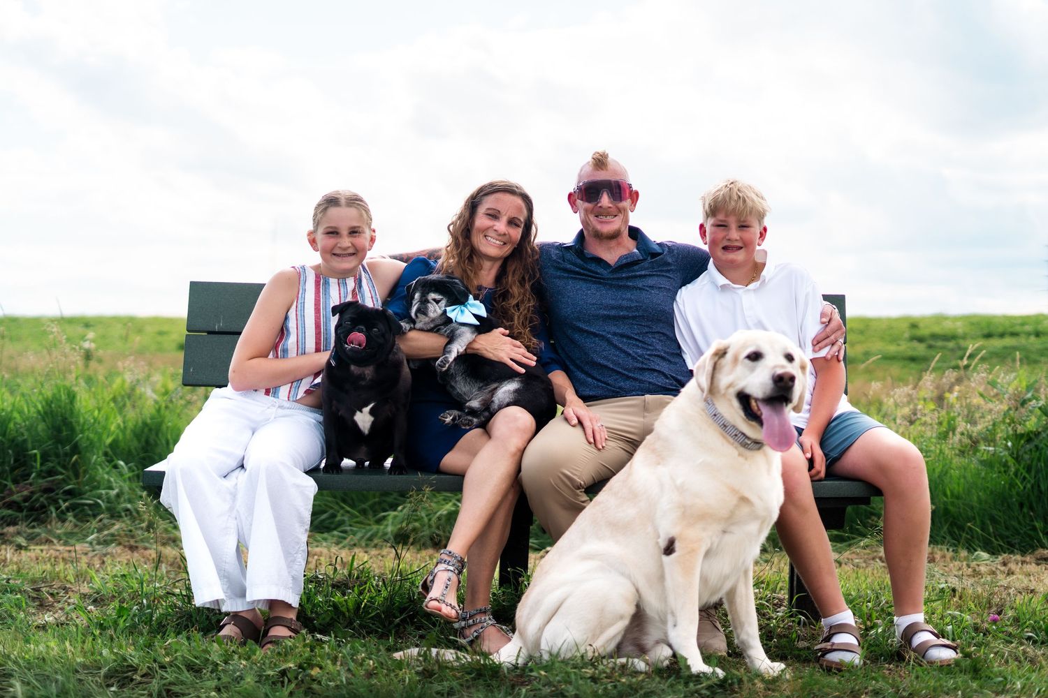 Family of four with two dogs sitting on a bench in a green field, smiling for a portrait outdoors.