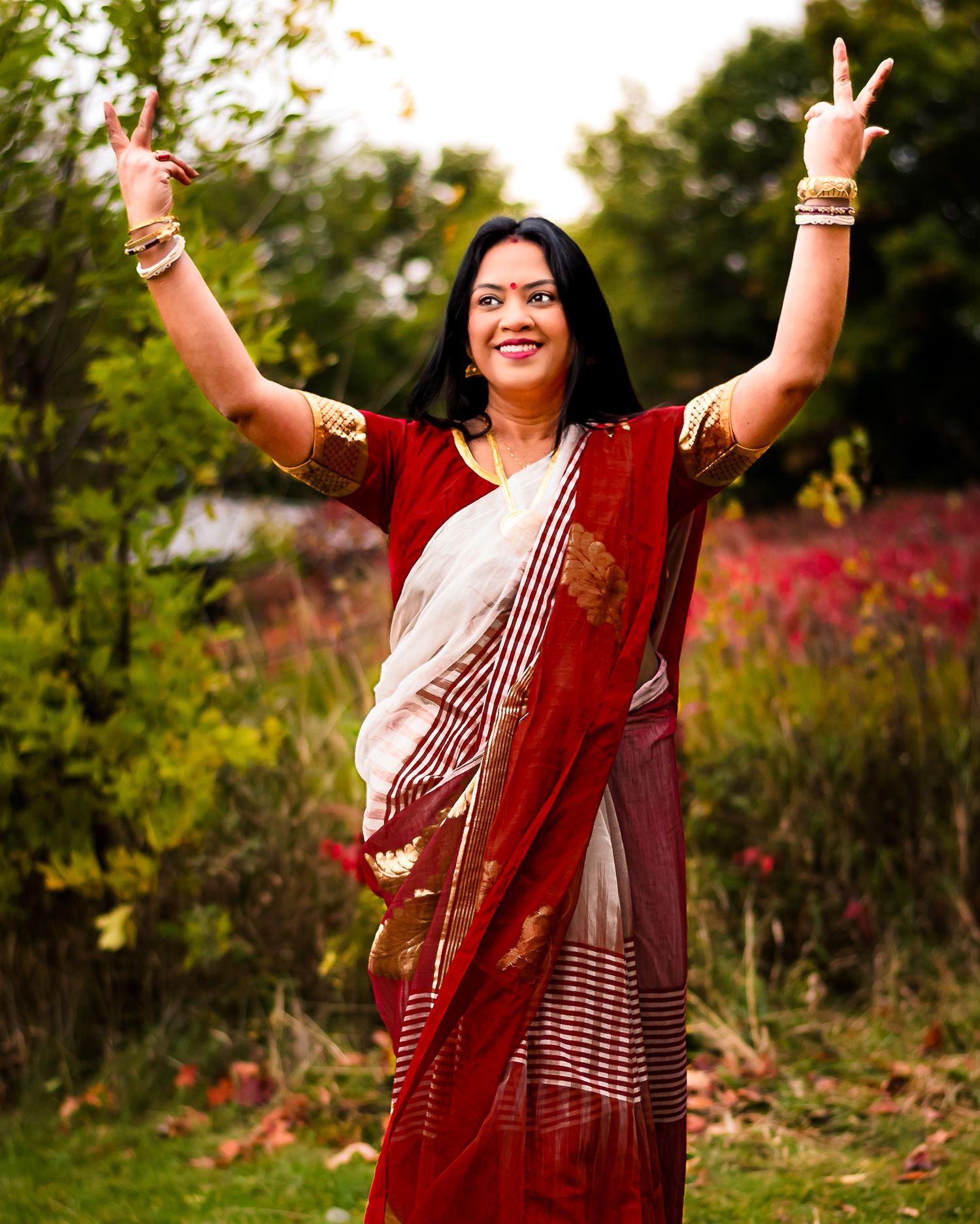 Woman in red and white saree with gold jewelry dancing outdoors with arms raised in a garden setting.