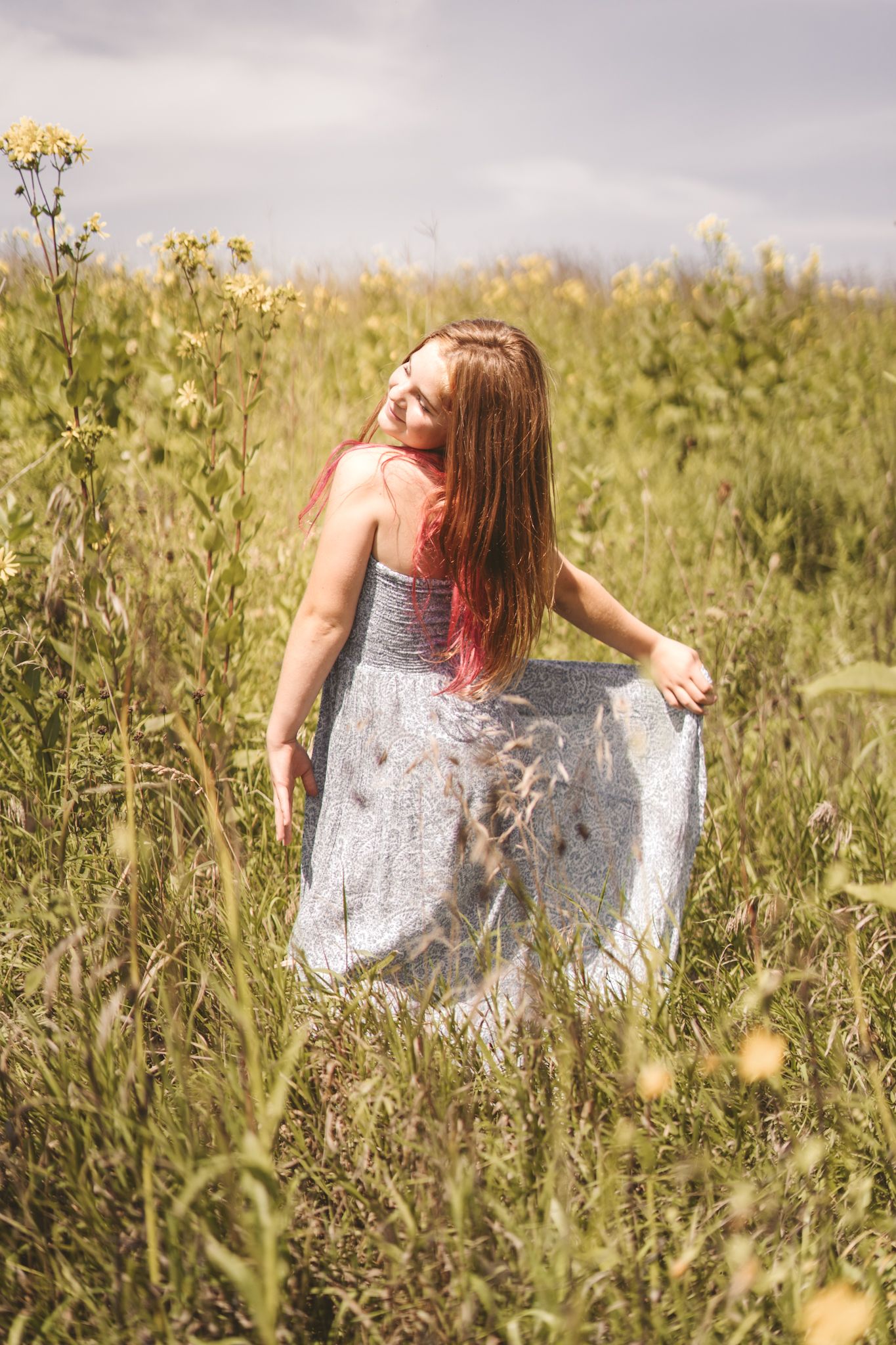 Young woman in gray dress holding skirt in wildflower meadow, long red hair flowing, smiling under cloudy sky.