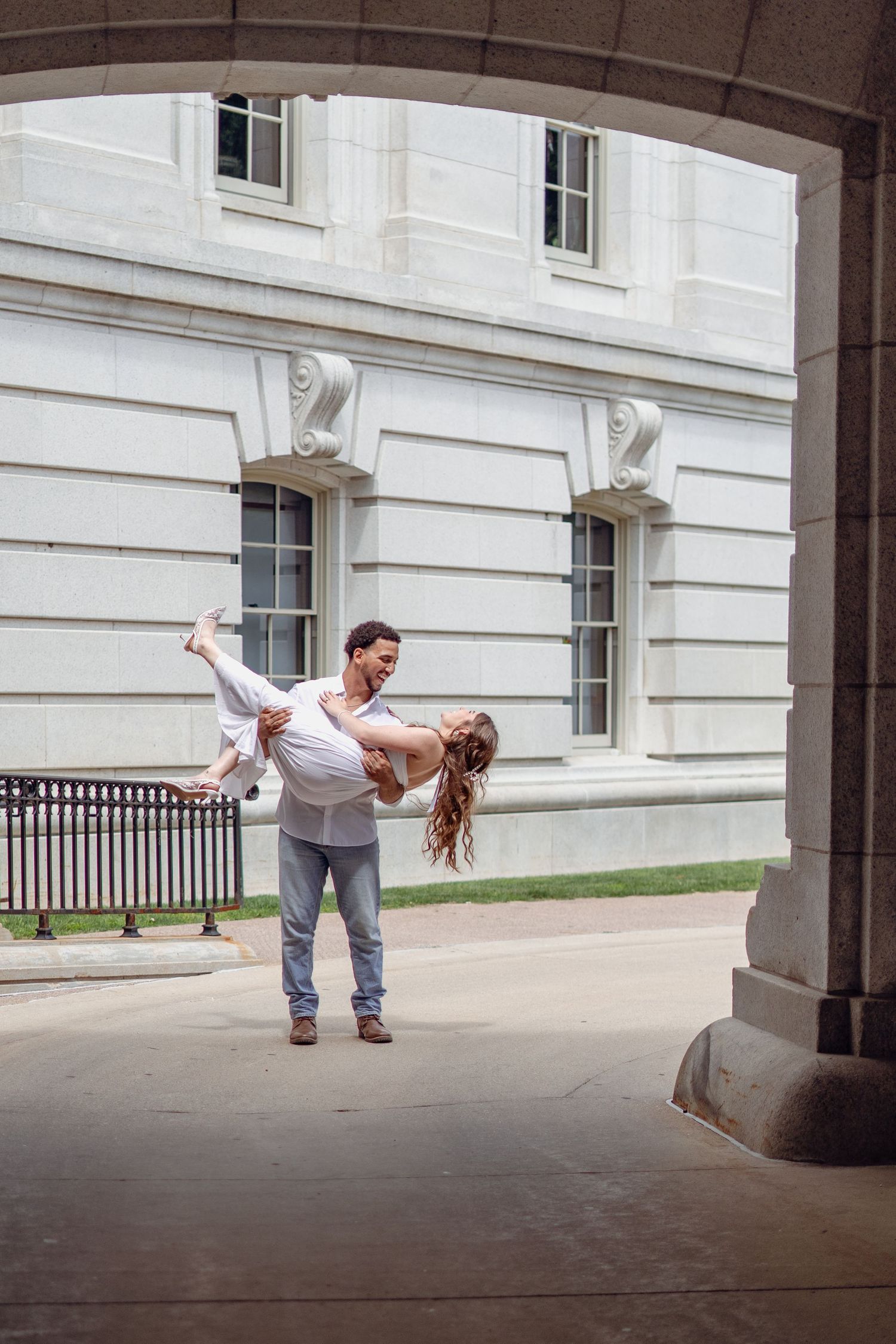Romantic couple dancing under stone arch, man lifting woman in white dress outside historic building.