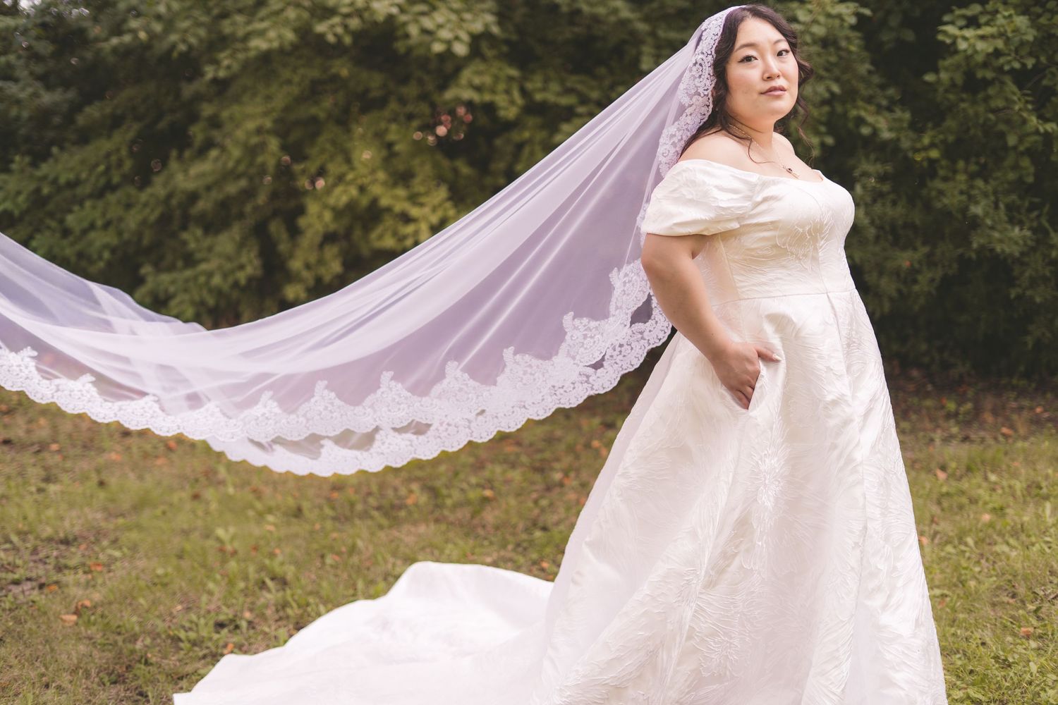 Plus-size bride in white off-shoulder wedding gown with flowing lace veil outdoors in green garden setting.