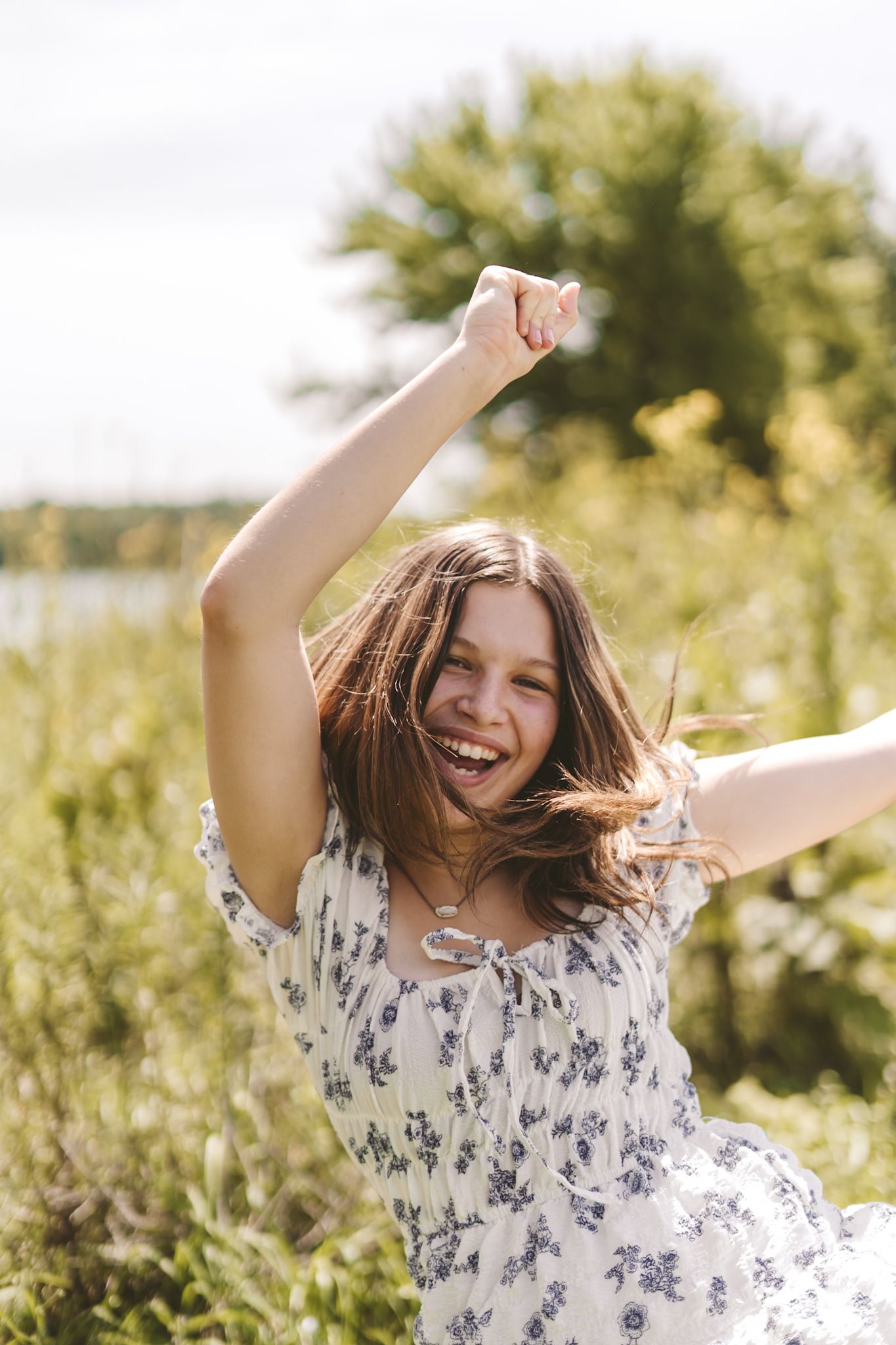 Happy young girl in floral dress dancing outdoors in sunny green meadow with arms raised, laughing joyfully.