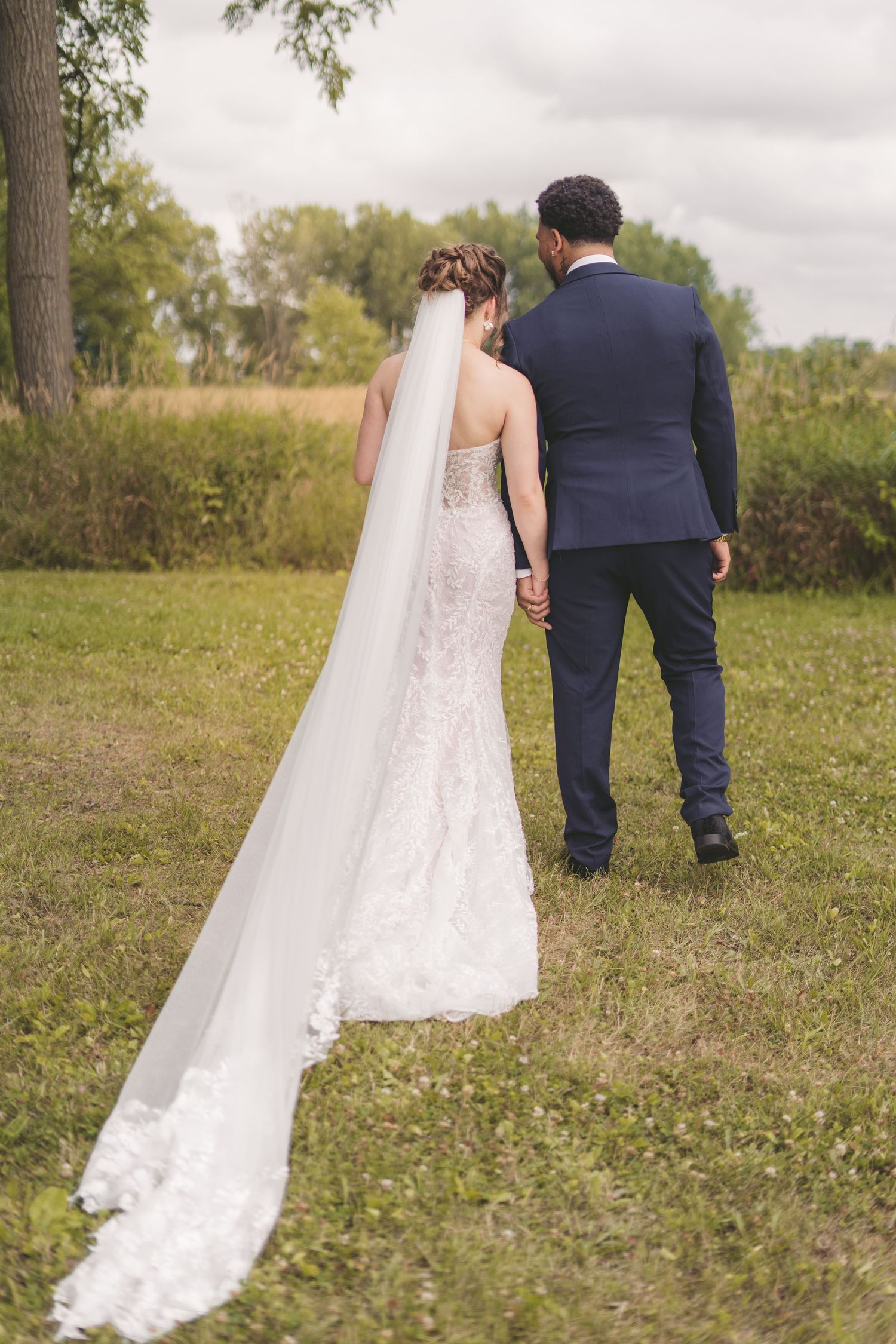 Bride and groom walking hand in hand outdoors, bride wearing lace gown with long flowing veil, groom in navy suit.