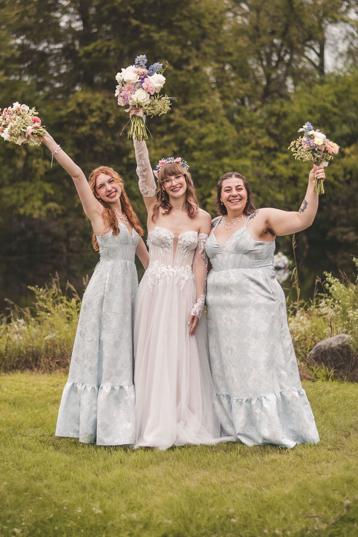 Bride in floral crown with two bridesmaids in silver gowns raising bouquets in celebration outdoors.