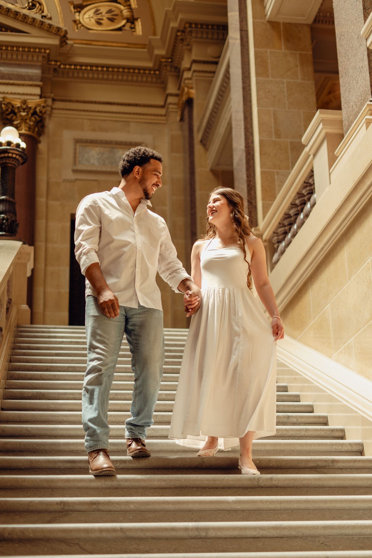 Happy couple holding hands walking down grand marble staircase, woman in white dress and man in white shirt and jeans.
