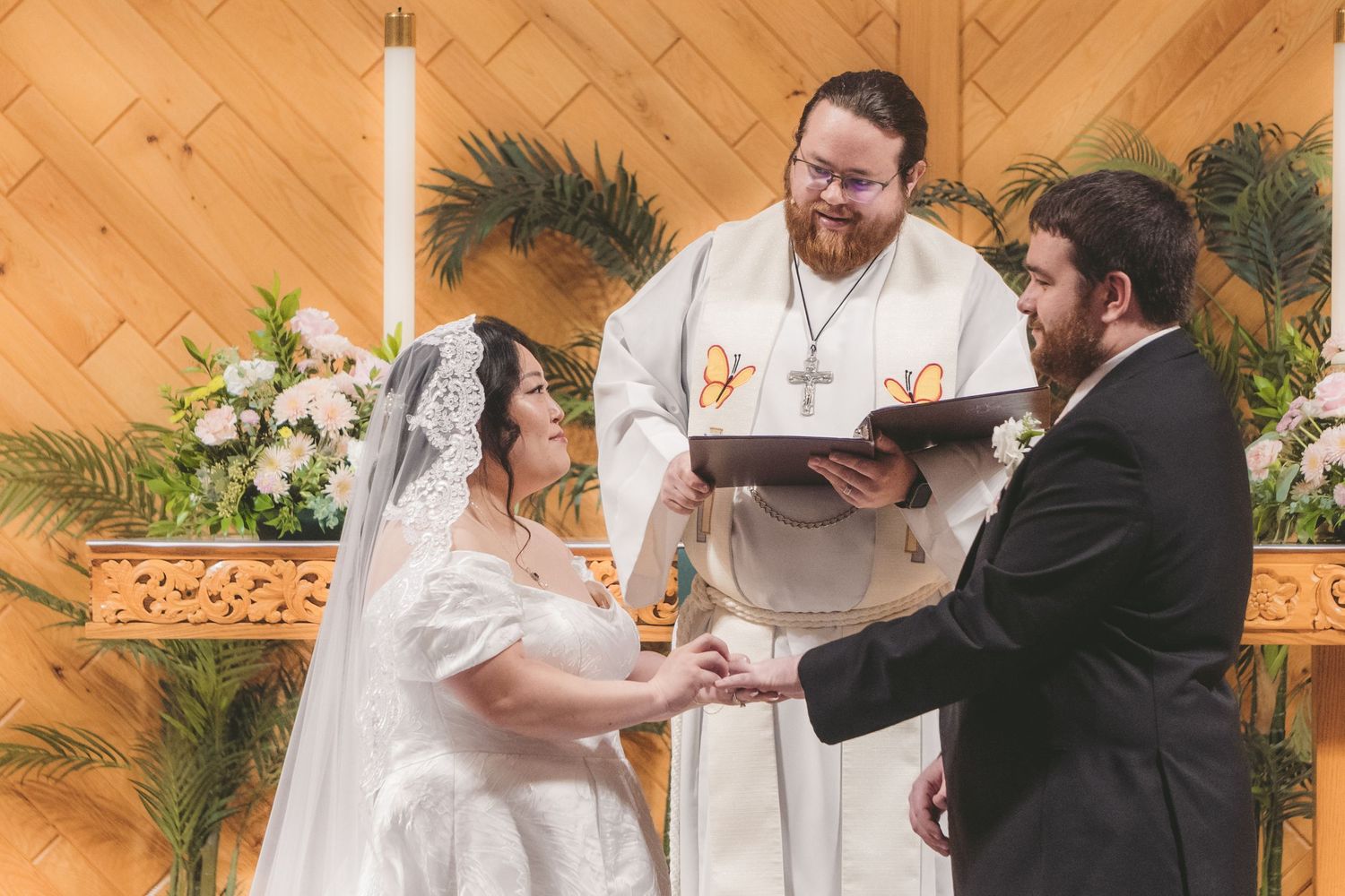 Bride and groom exchange vows at altar with officiant holding book during wedding ceremony in decorated chapel.