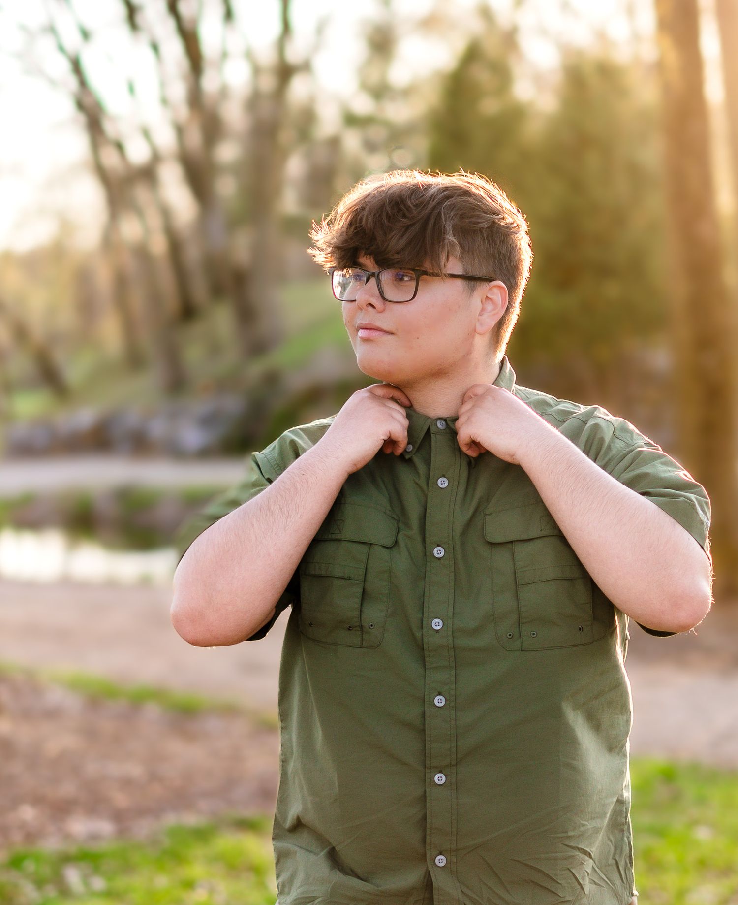 A person in glasses and olive green button-up shirt poses outdoors during golden hour.