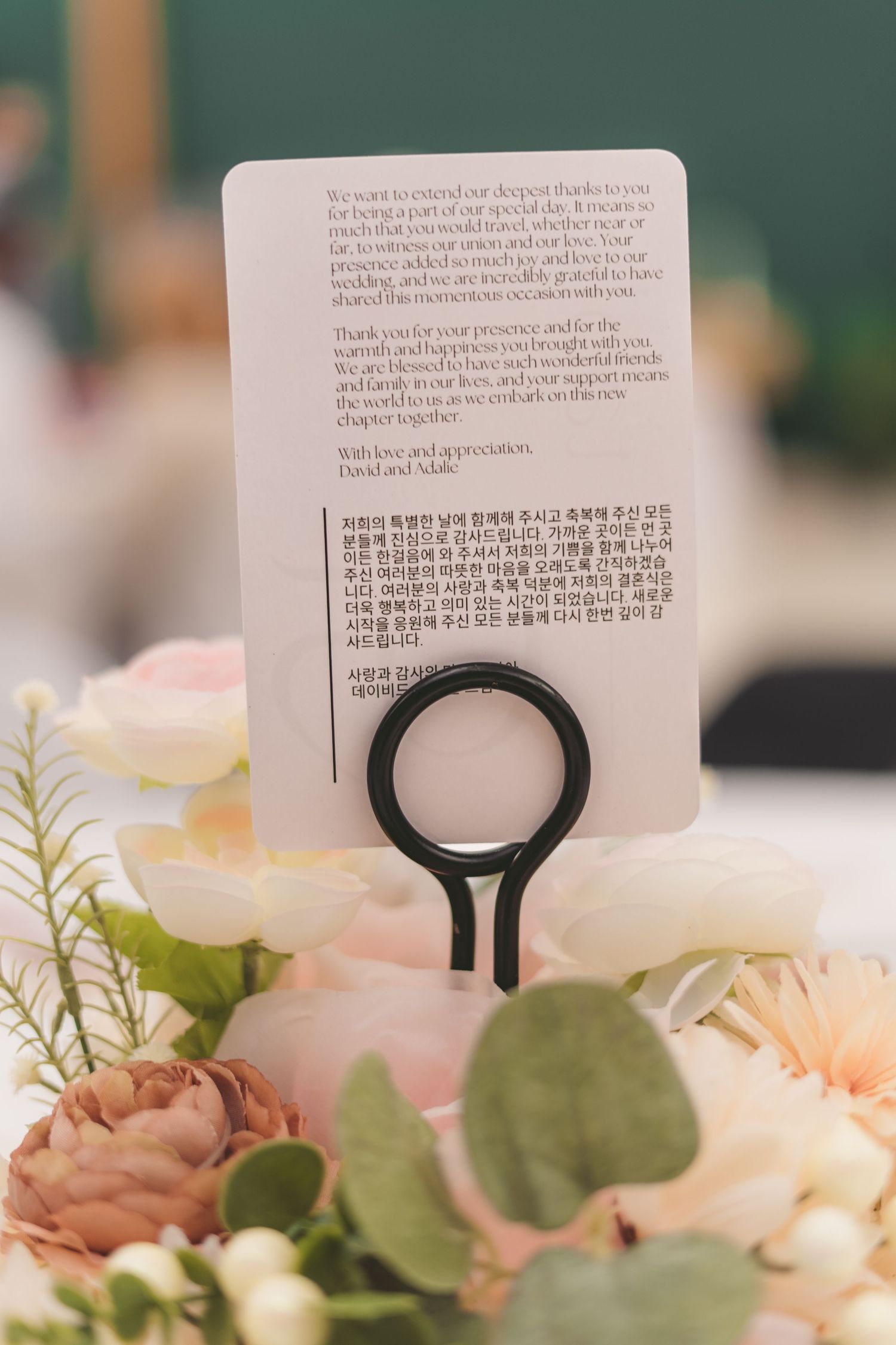 Wedding table card with bilingual English and Korean text displayed in a black ring holder among pink and white floral arrangements.