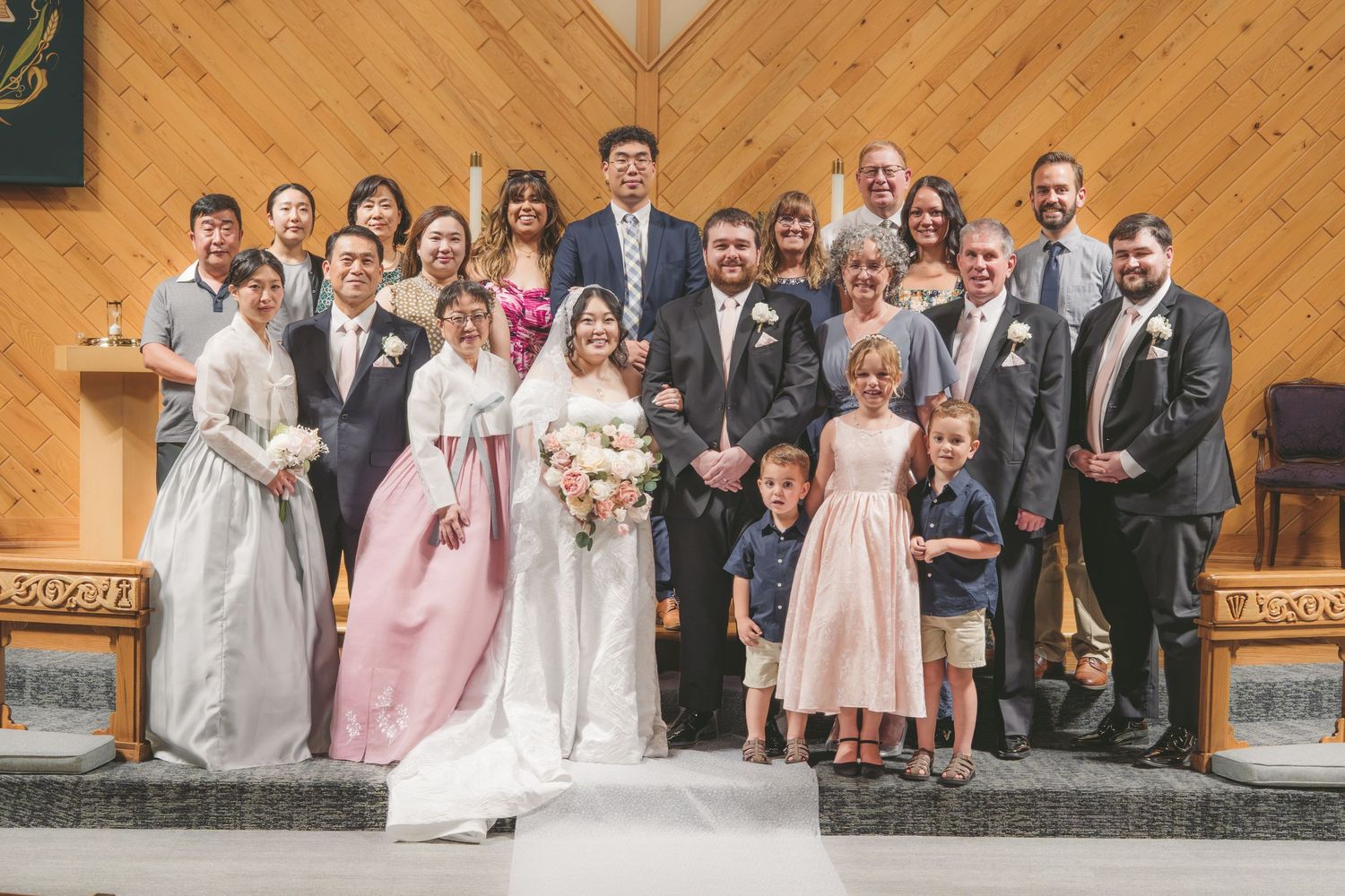 Wedding party poses together in a church, bride in white gown, guests in formal attire, children in front row.