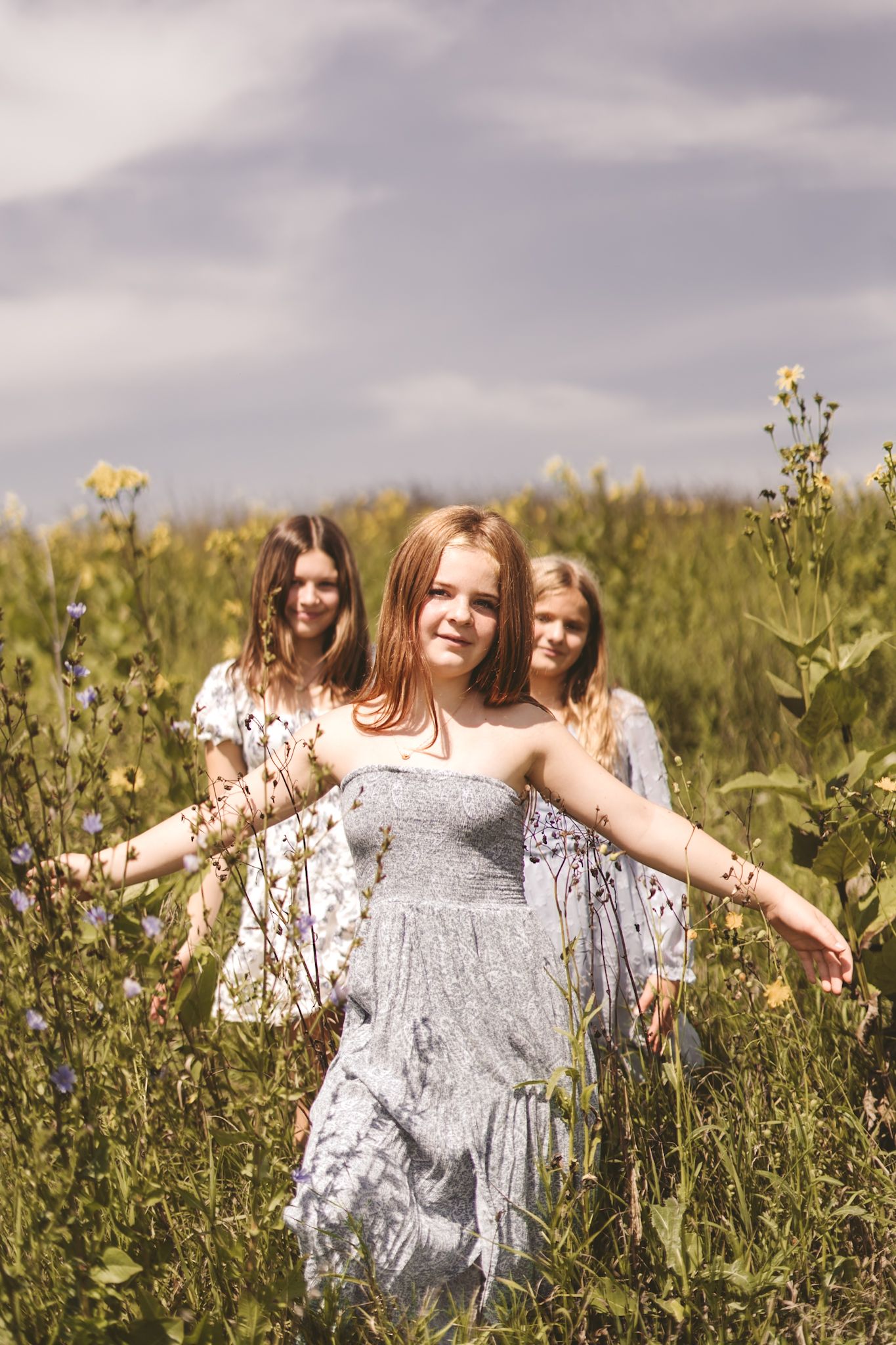 Three young women in floral dresses walk through a wildflower meadow under a cloudy sky.