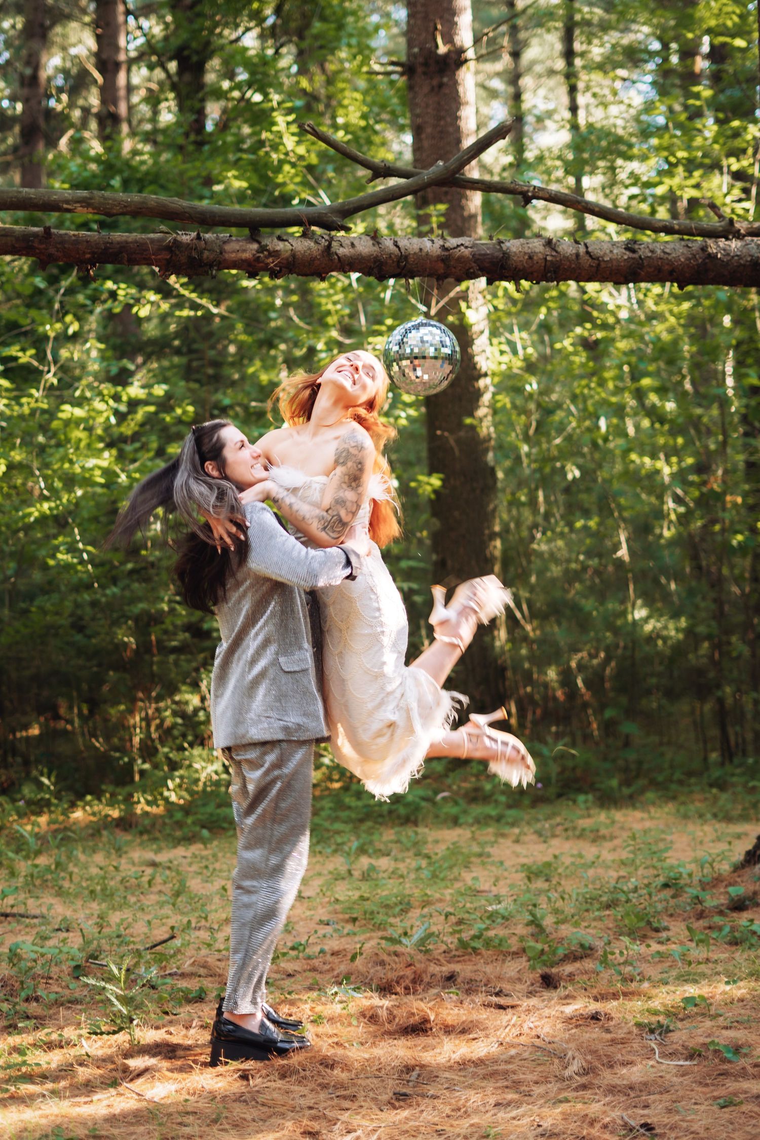 Couple celebrating wedding in forest, groom lifting bride in white dress near disco ball hanging from tree branch.