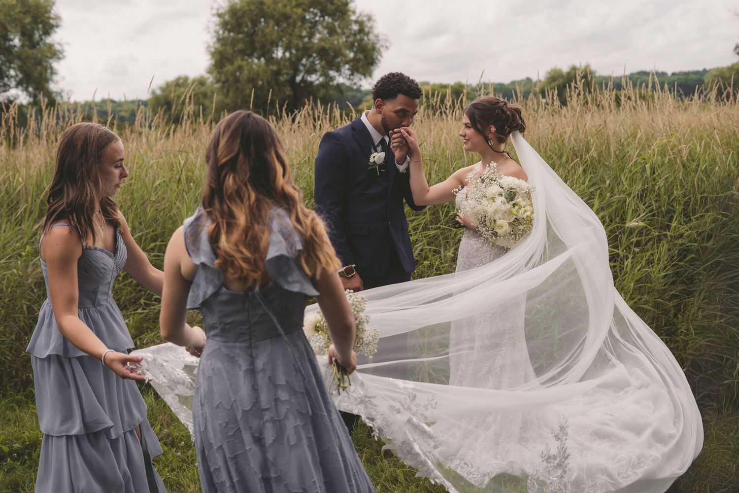 Bride and groom share a tender moment as bridesmaids in gray dresses hold a flowing veil outdoors.