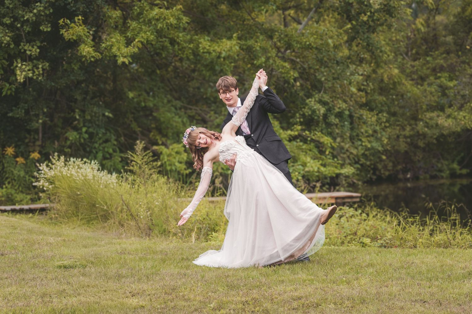 Groom in black suit dips bride in white flowing gown during outdoor wedding dance on green lawn.