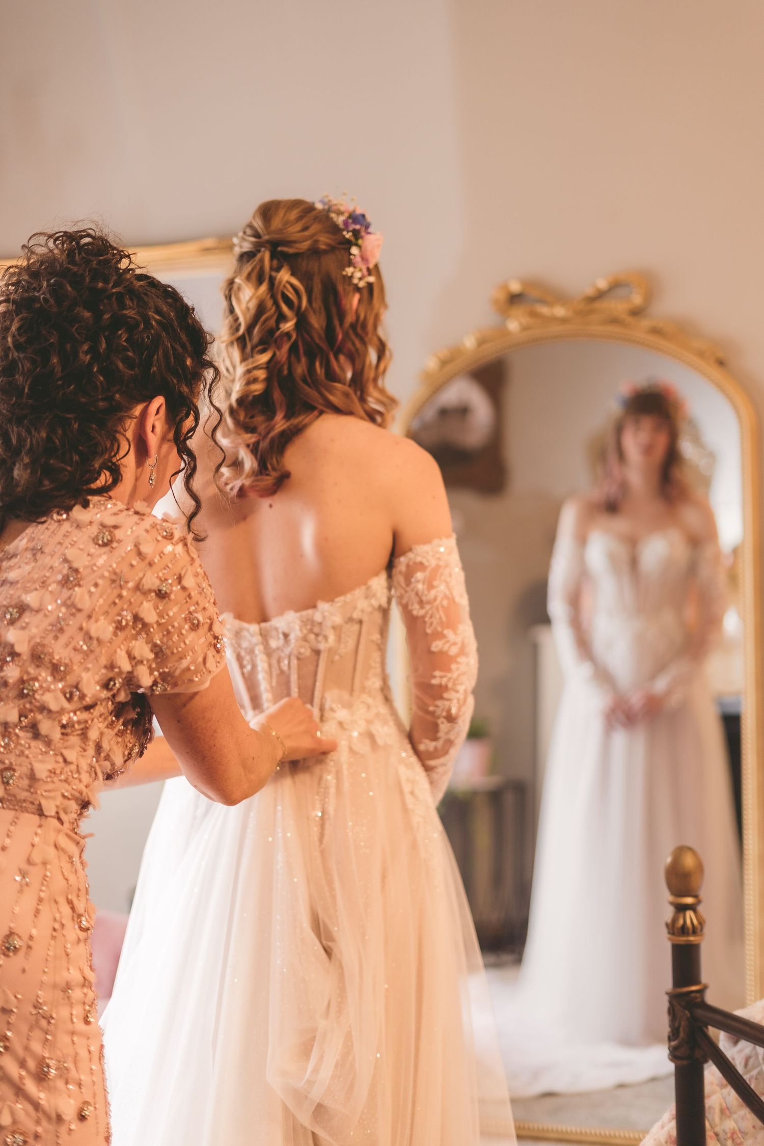 Bride getting her corset wedding dress laced up by bridesmaid, reflected in ornate gold mirror.