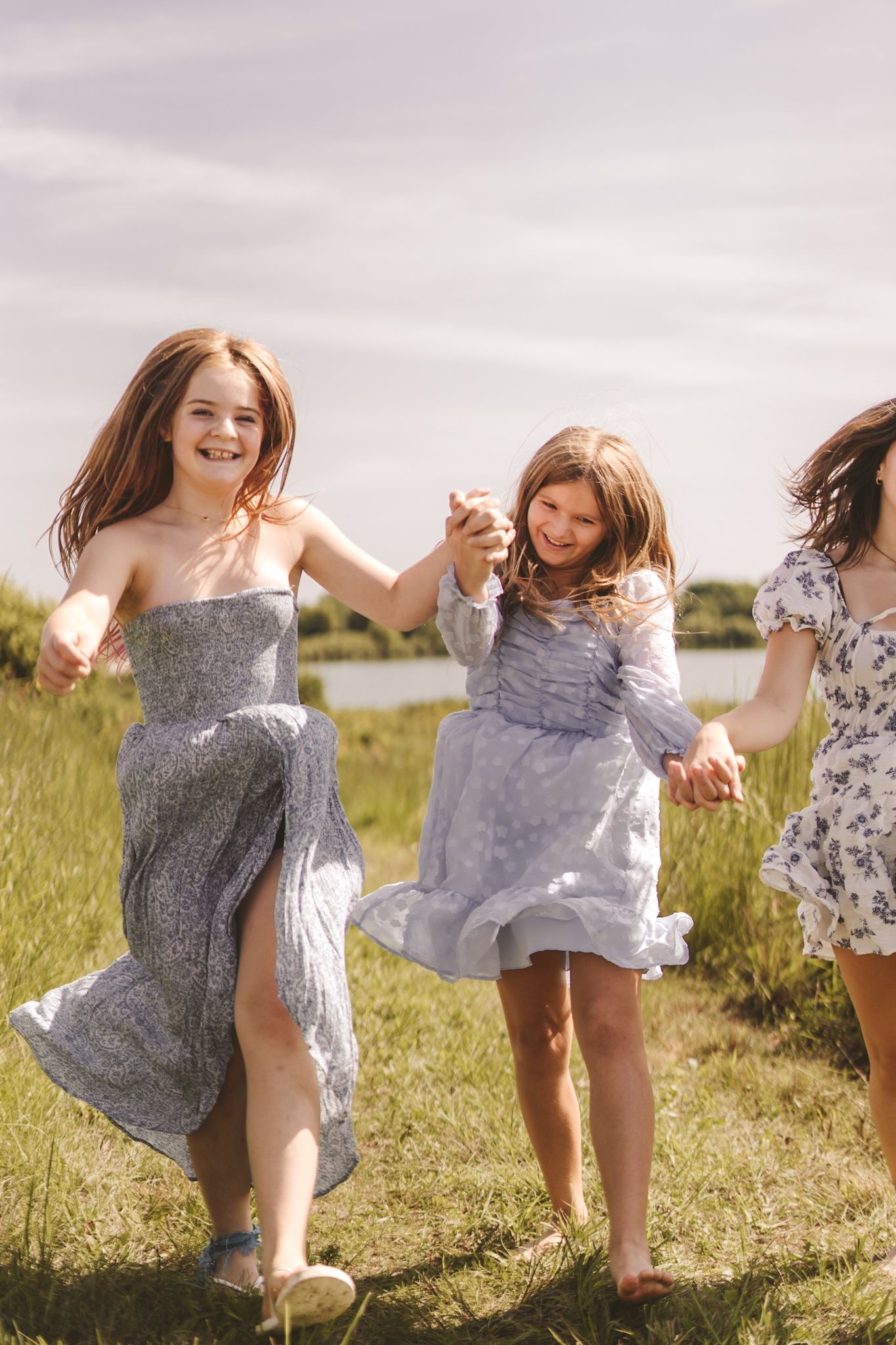 Three young girls in flowy dresses holding hands and running through a sunny green field near a lake.