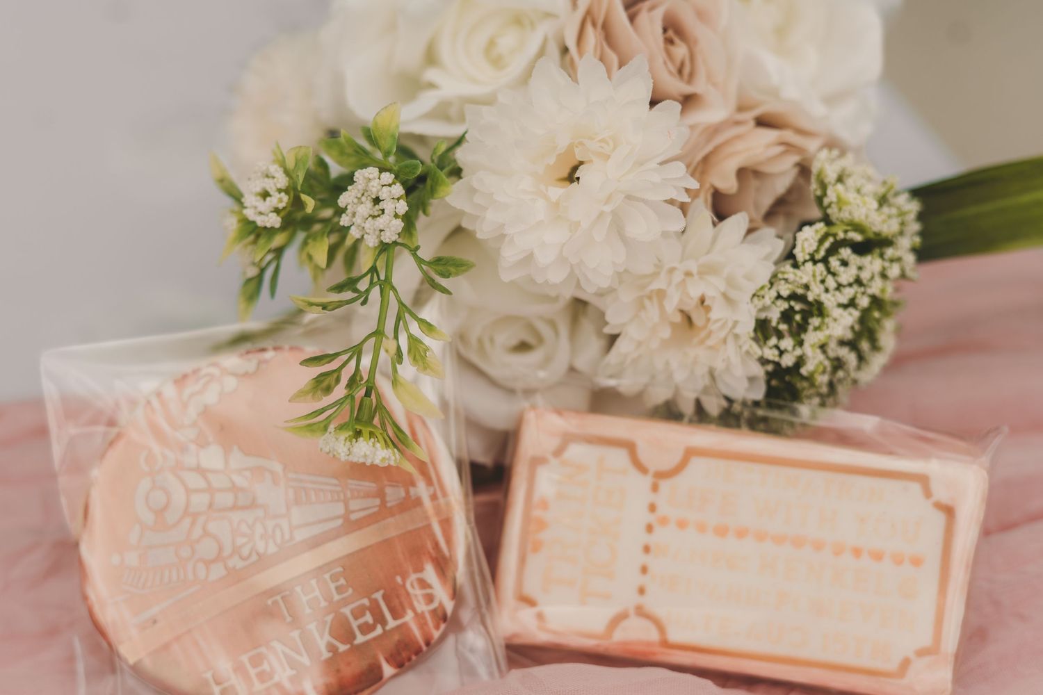 Elegant wedding bouquet of white dahlias and roses beside peach-colored favor cookies in clear packaging.