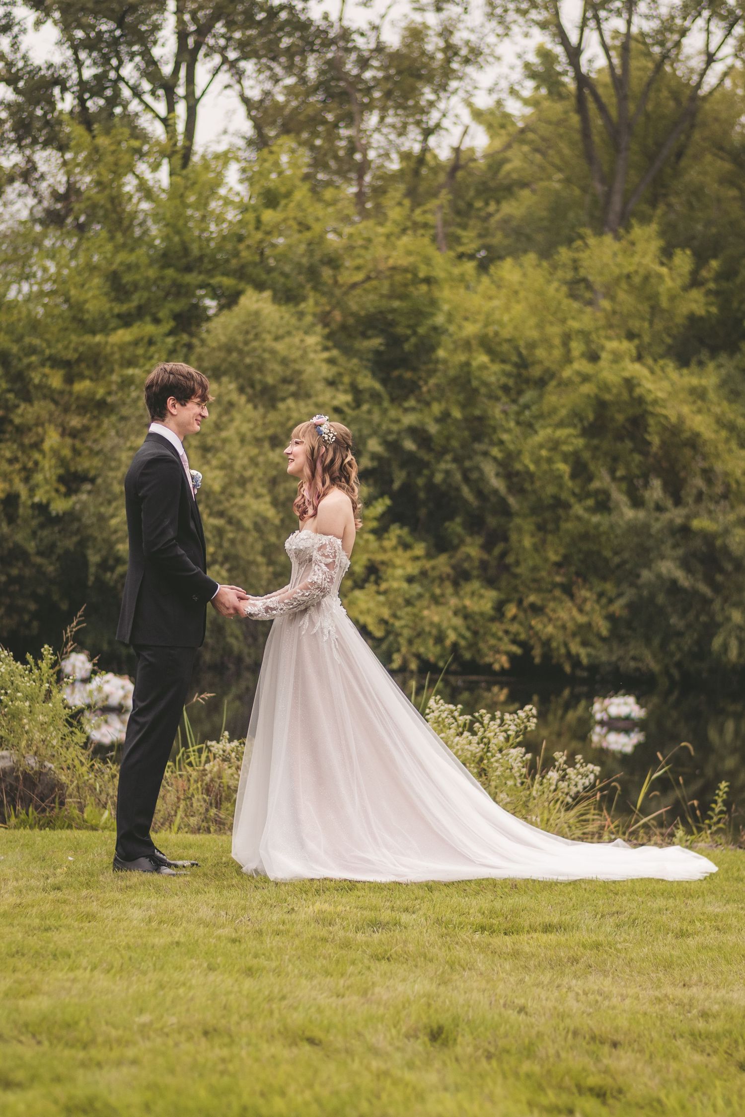 A bride and groom hold hands in a scenic garden, her flowing lace gown trailing on the grass.