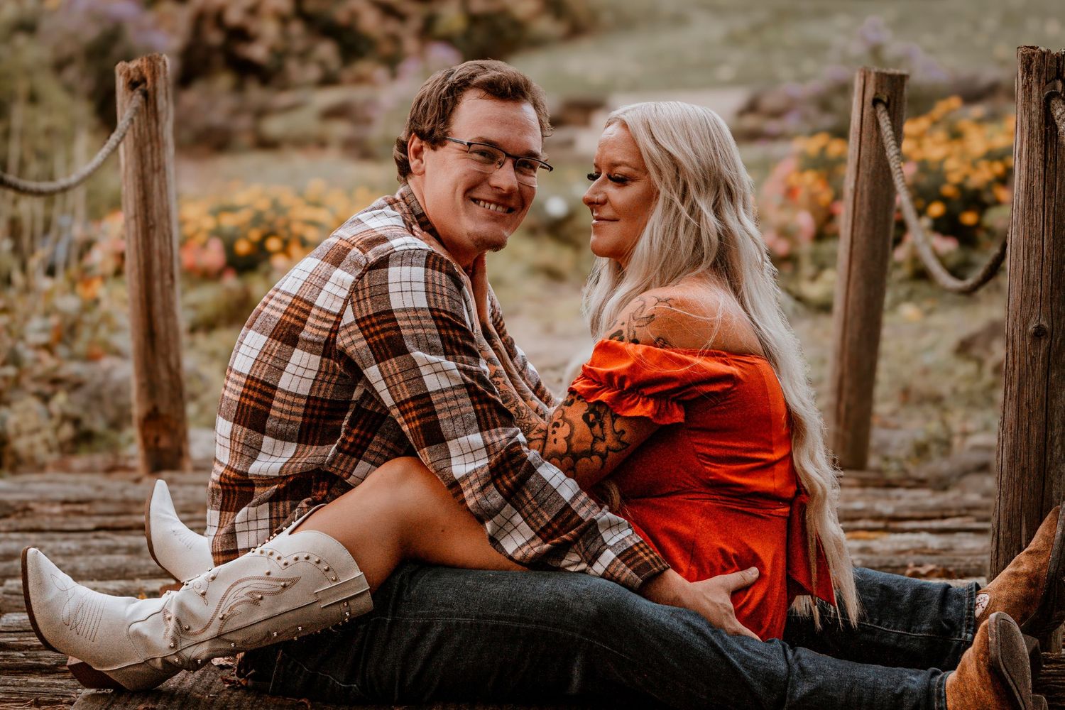 Couple in plaid shirt and red dress share tender moment on rustic wooden bridge.