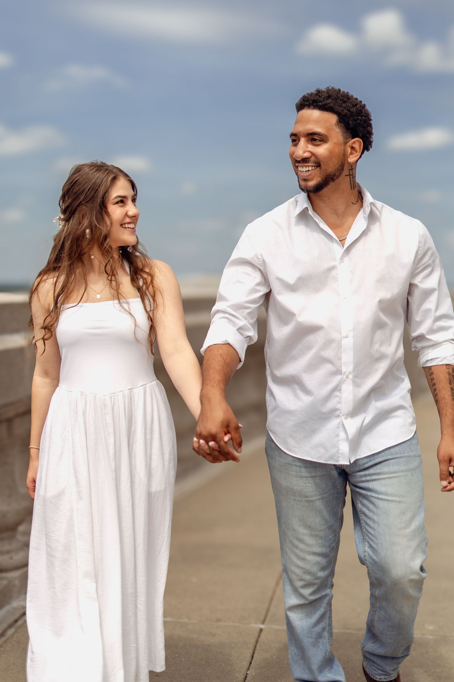 Smiling interracial couple holding hands walking outdoors, man in white shirt and jeans, woman in white sundress.