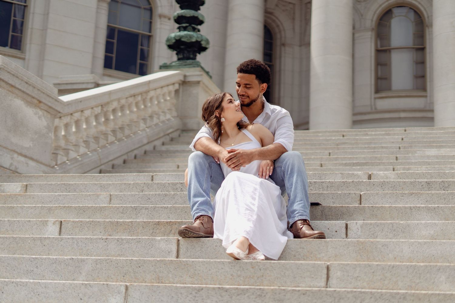 Couple sitting on marble steps of a grand building, embracing romantically in casual and white dress attire.