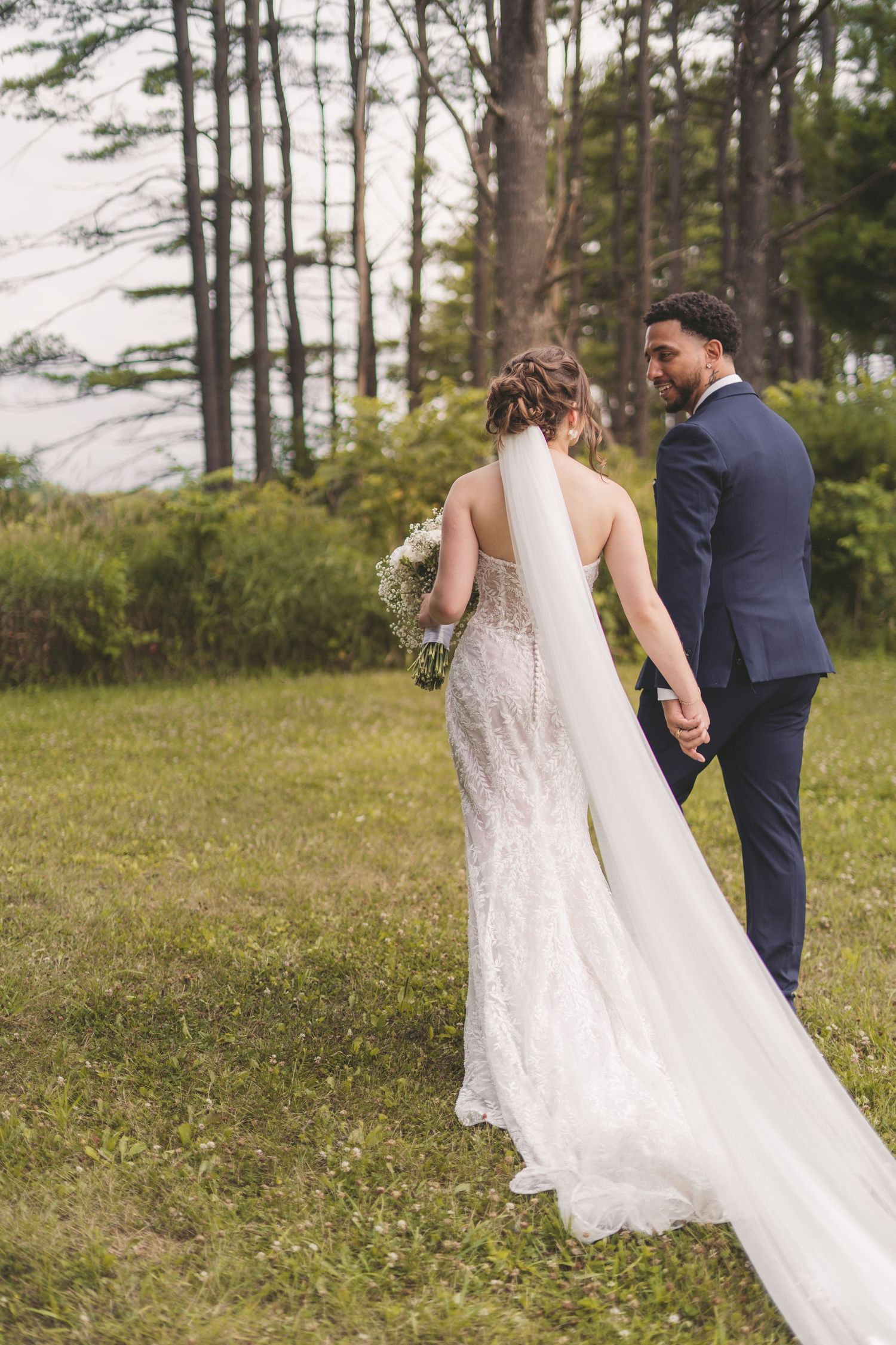 Bride and groom walking hand in hand through lush green forest, bride wearing lace gown with long veil.