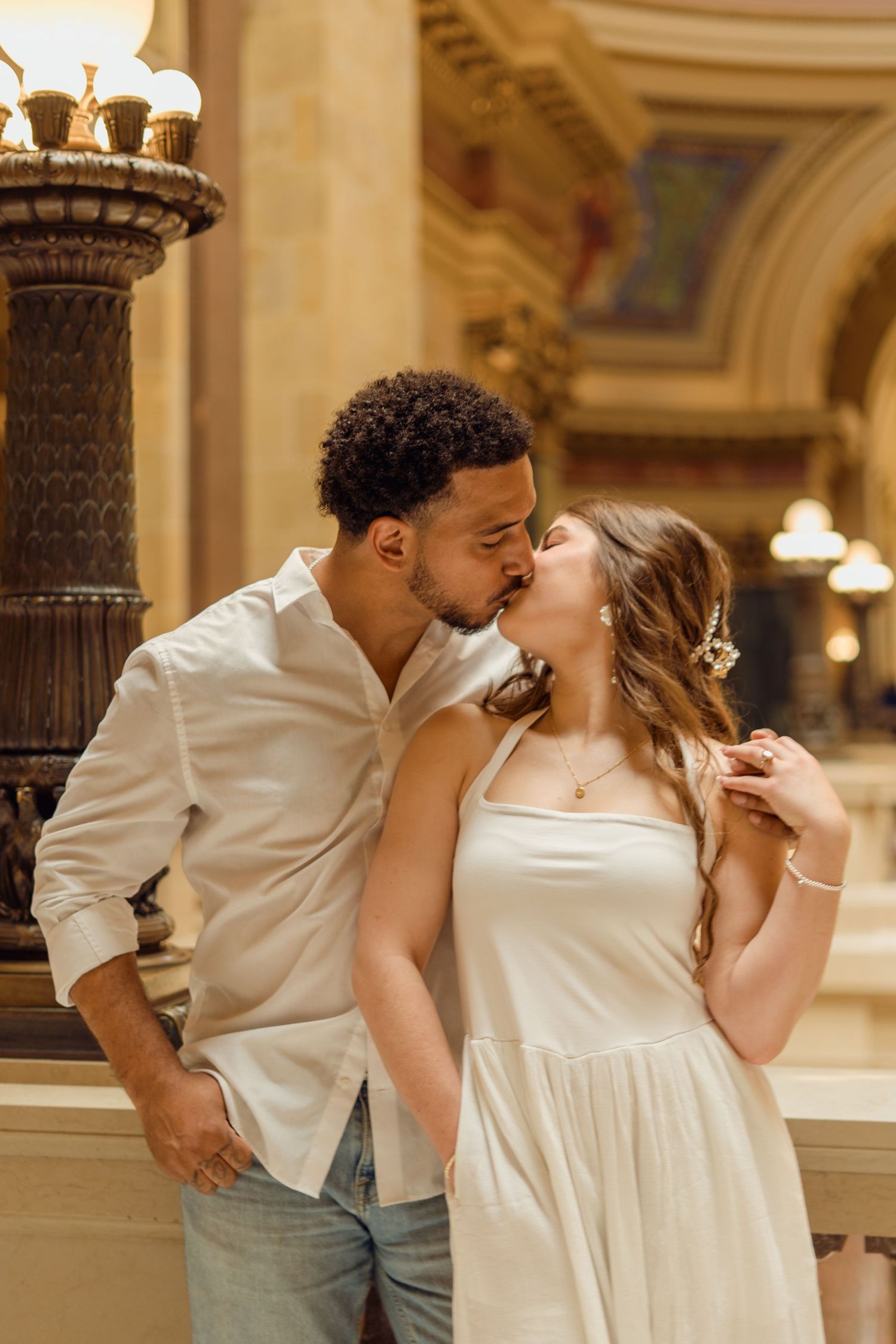 Interracial couple sharing a romantic kiss inside an elegant building with ornate architecture.