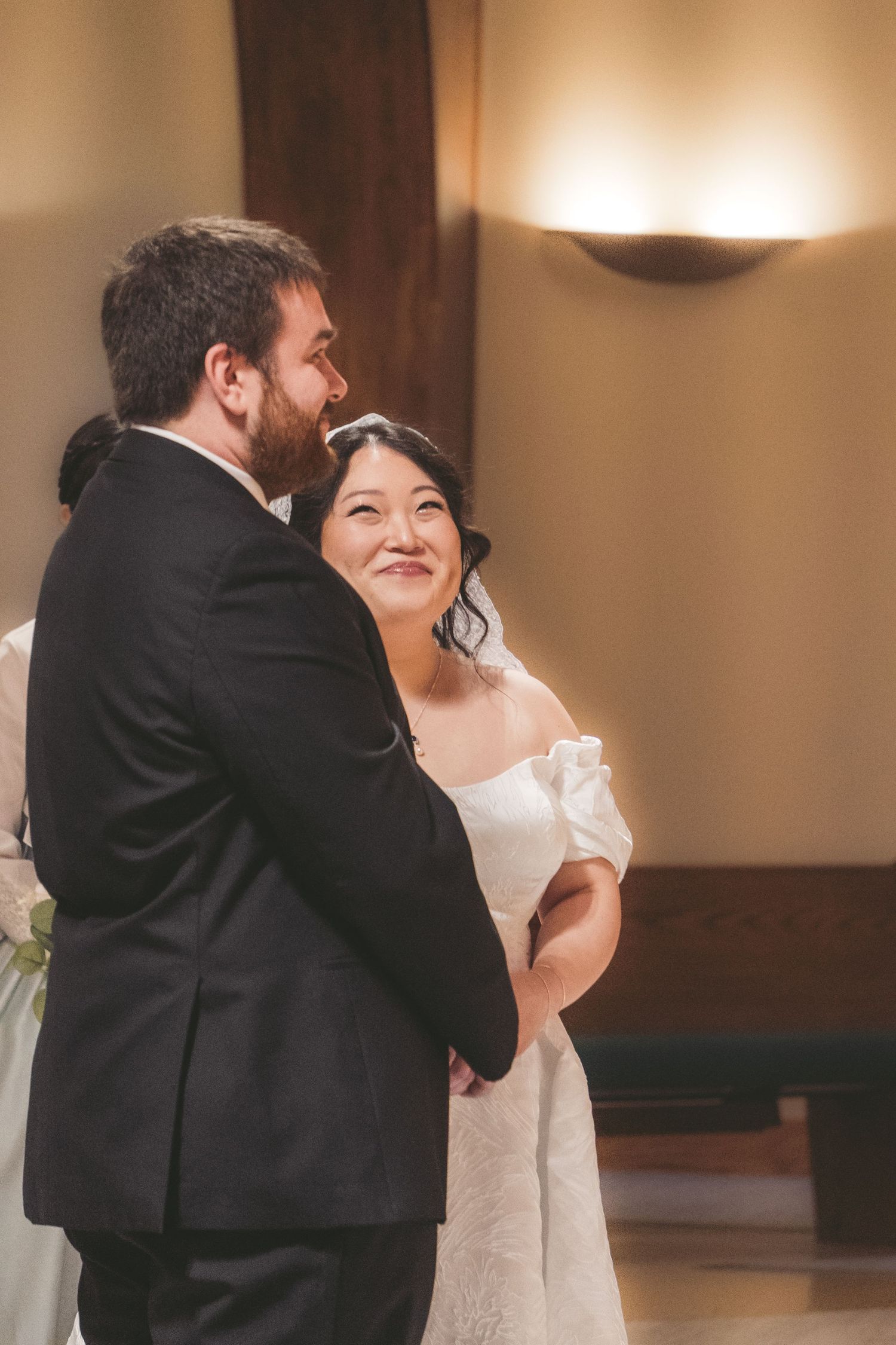 Bride in white off-shoulder gown smiles at groom in black suit during wedding ceremony in elegant venue.